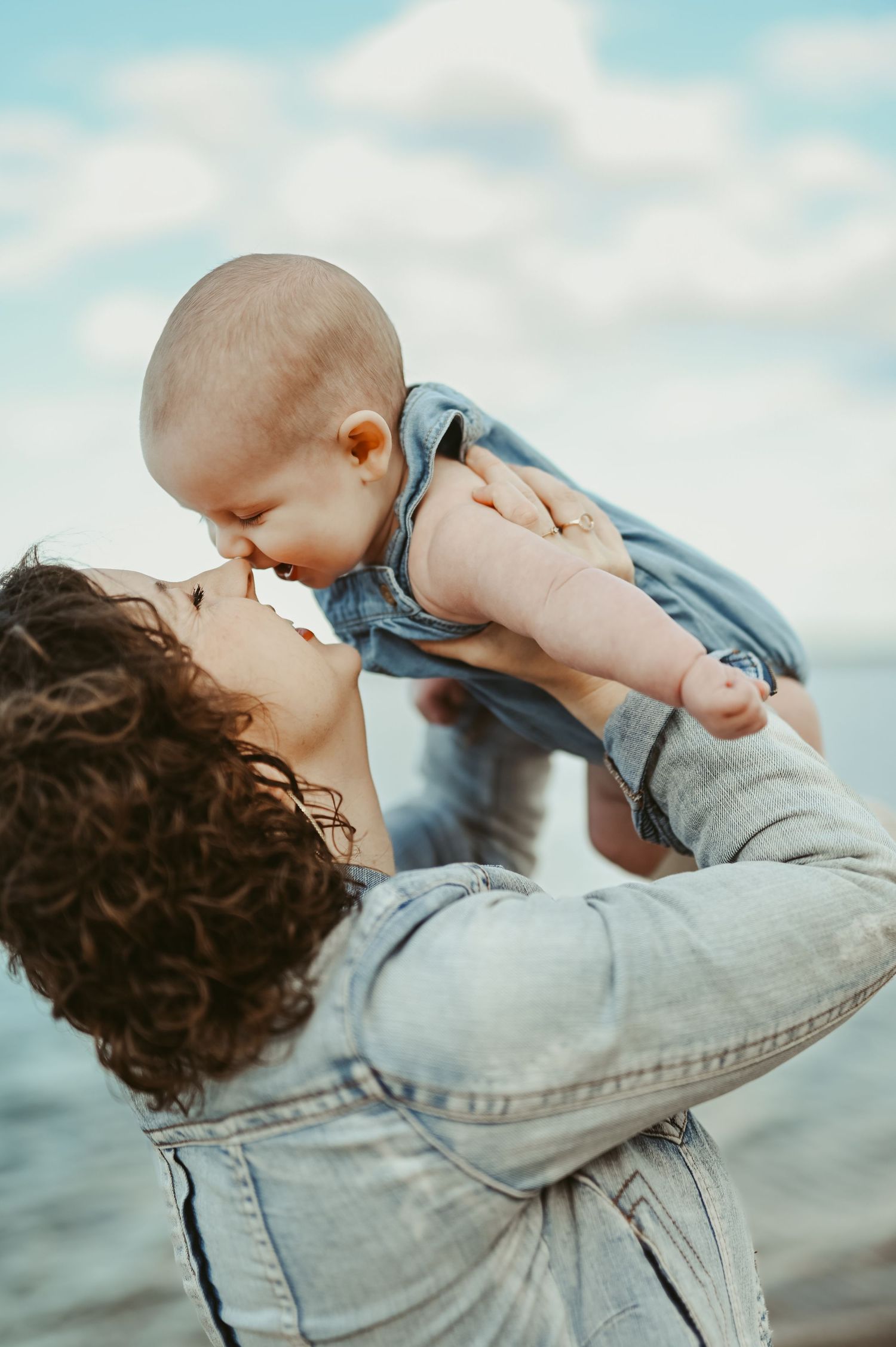 A curly-haired mother in a denim jacket lifts her smiling baby up to kiss them near a serene waterfront.