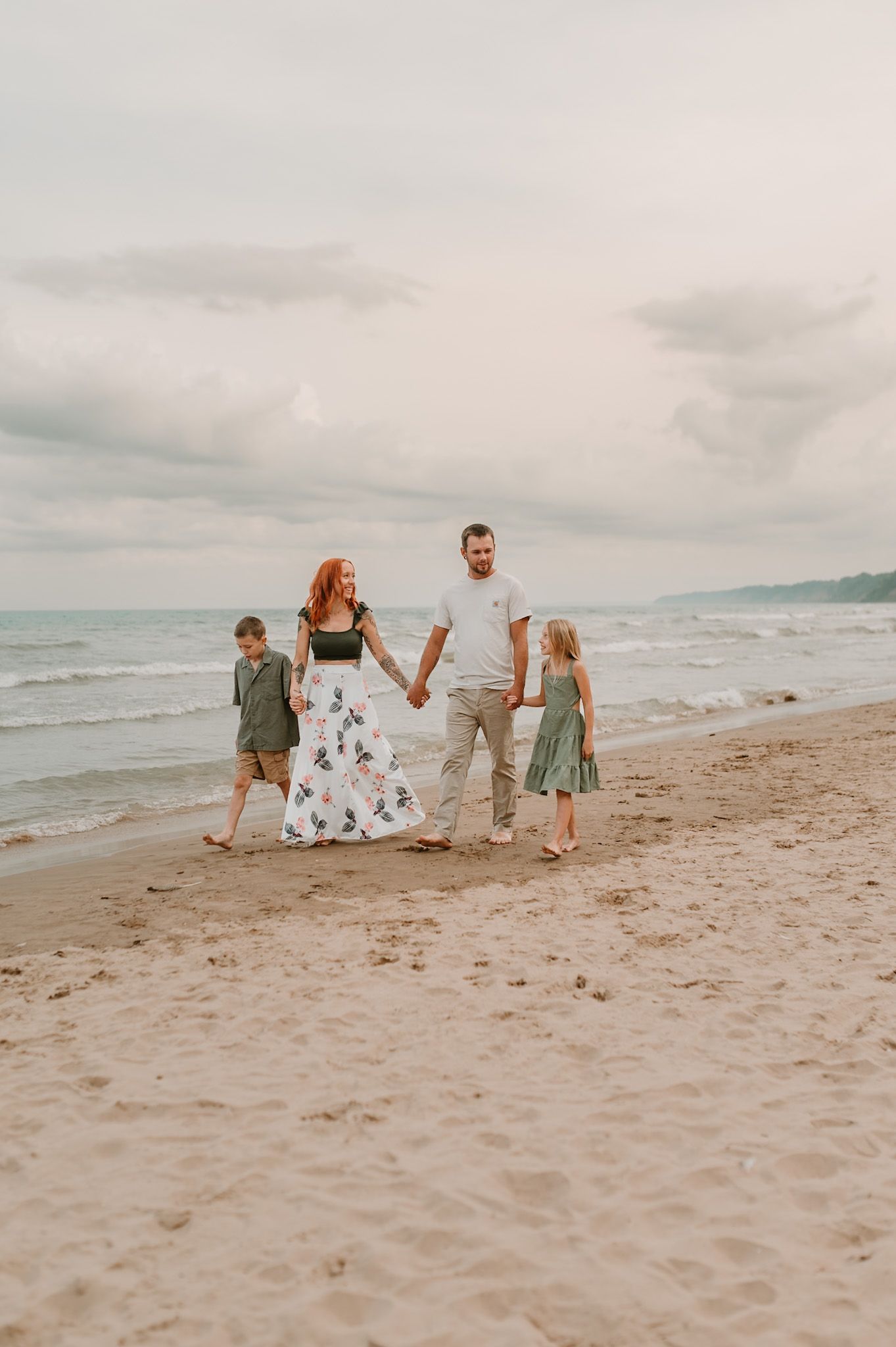 Family of four holding hands walking on a sandy beach with ocean waves in the background.