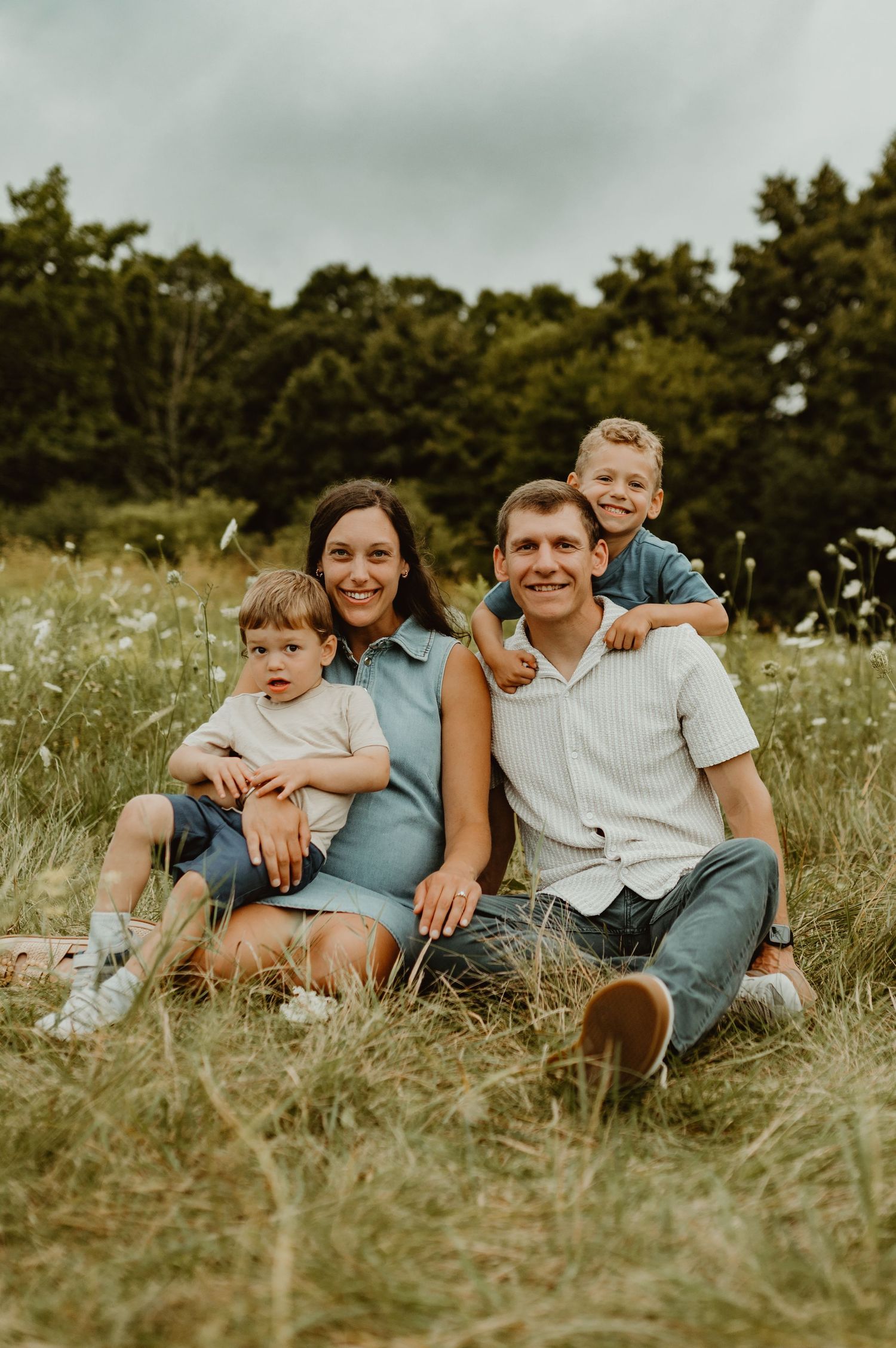 Happy family of four sitting in a wildflower meadow, smiling outdoors with lush green trees in the background.