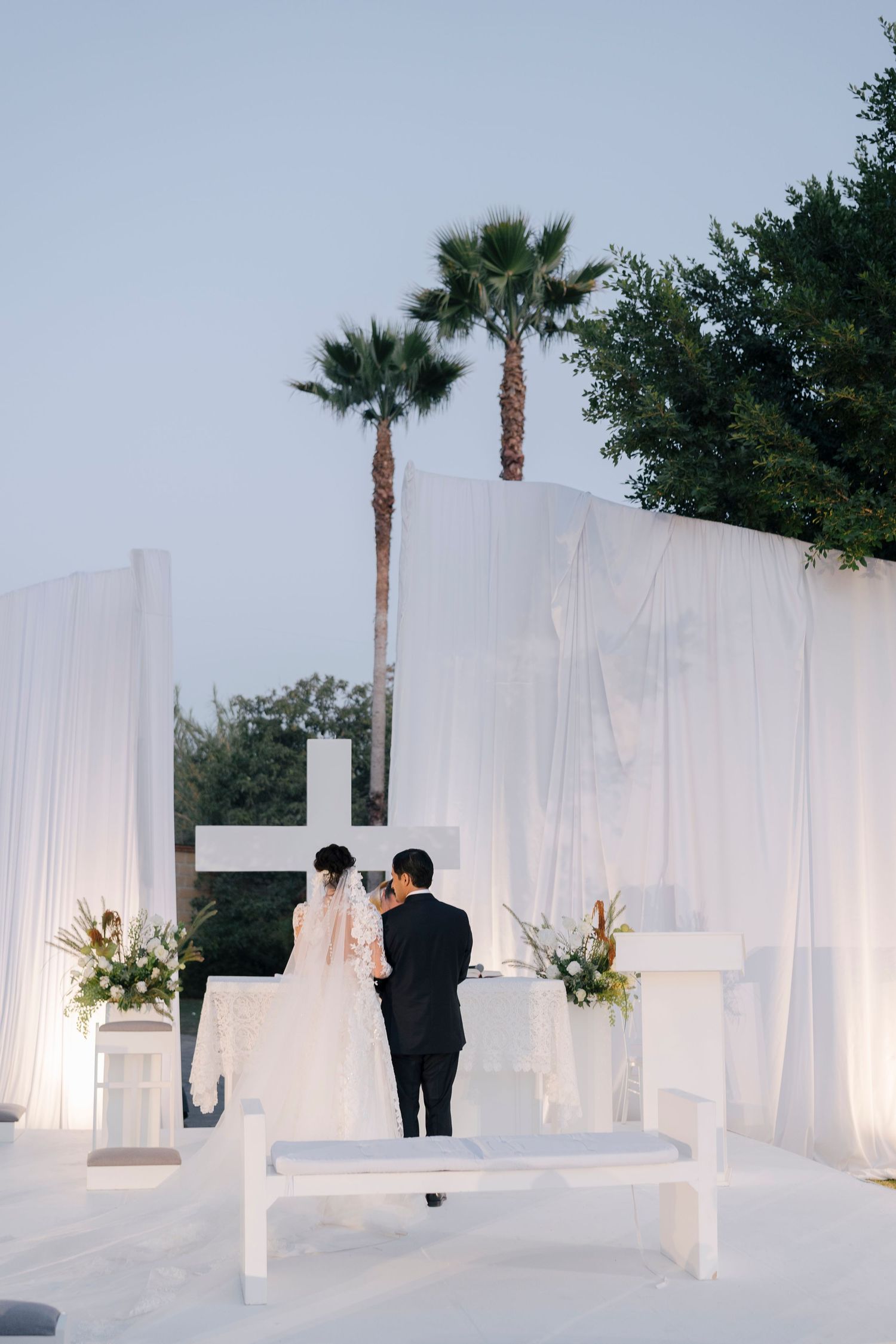 Bride and groom share a kiss at elegant outdoor wedding ceremony with white draping, cross altar, and palm trees at dusk.