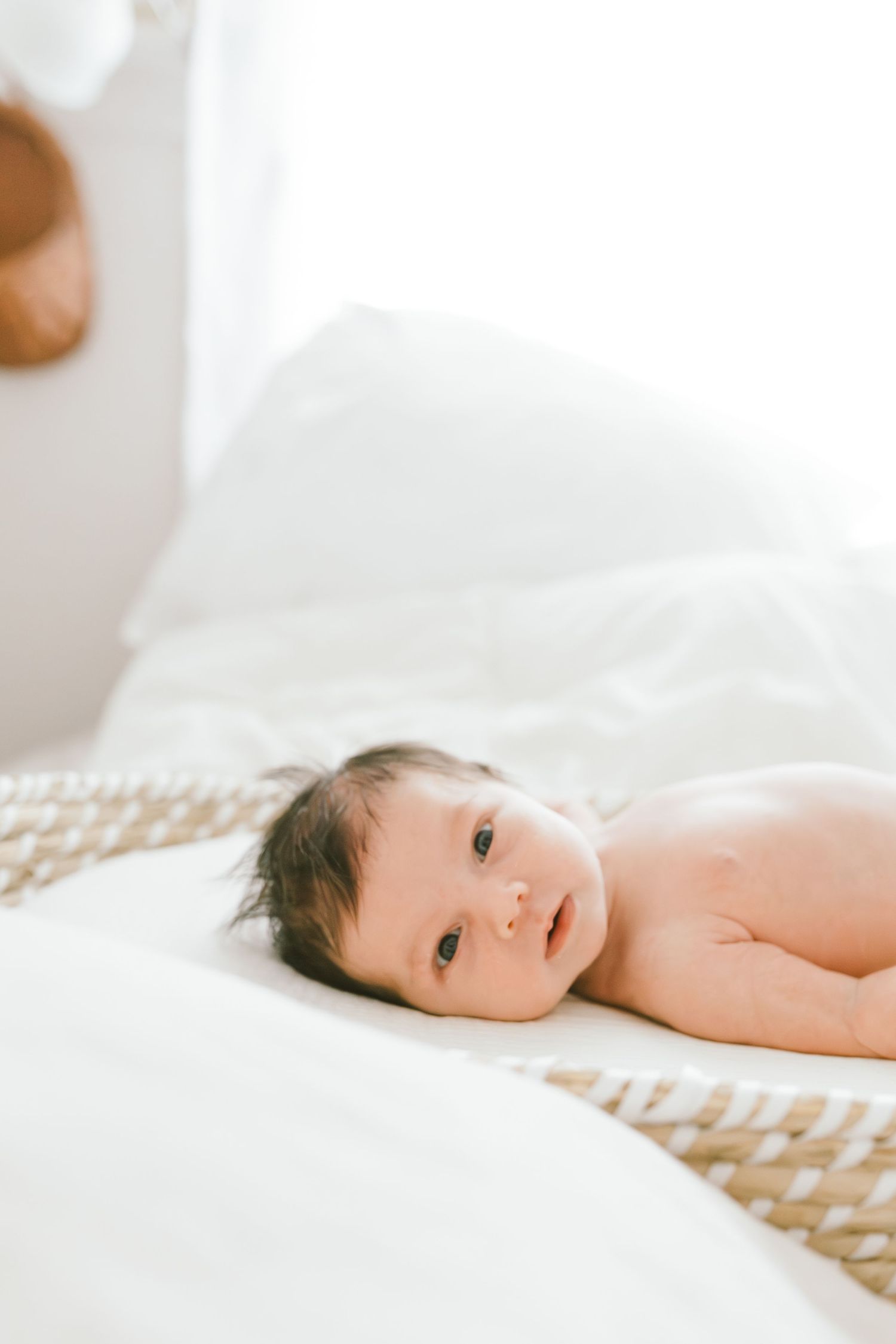 A newborn baby with dark hair lying on white bedding, gazing softly at the camera in a bright, airy room.
