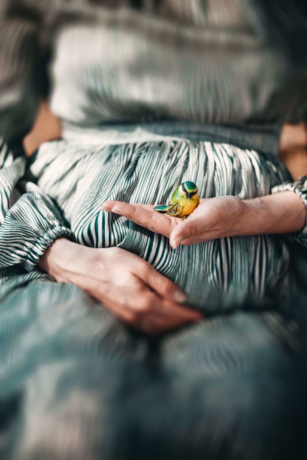 A small colorful bird perched on a woman's hand, wearing a striped green dress, in a serene close-up shot.