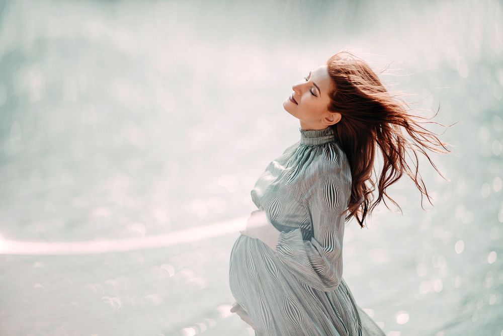 A pregnant woman in a flowing blue dress with windswept hair poses gracefully against a soft, dreamy background.