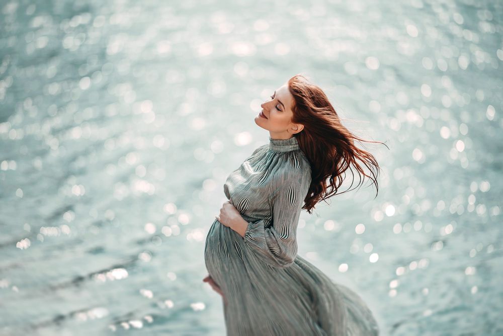 Pregnant woman in flowing teal dress poses gracefully by sparkling water with hair blowing in the wind.
