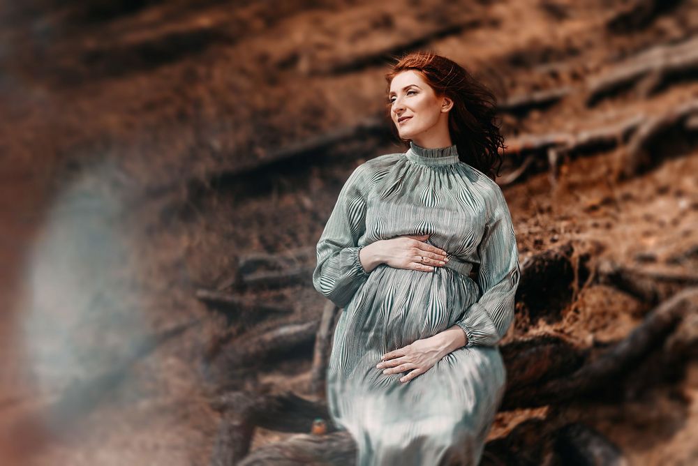 Smiling pregnant woman in teal dress cradles baby bump outdoors against rustic brown rocky background.