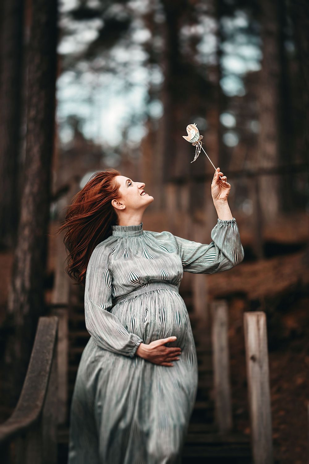 Pregnant redhead woman in teal dress holds white rose aloft outdoors near rustic wooden structure.