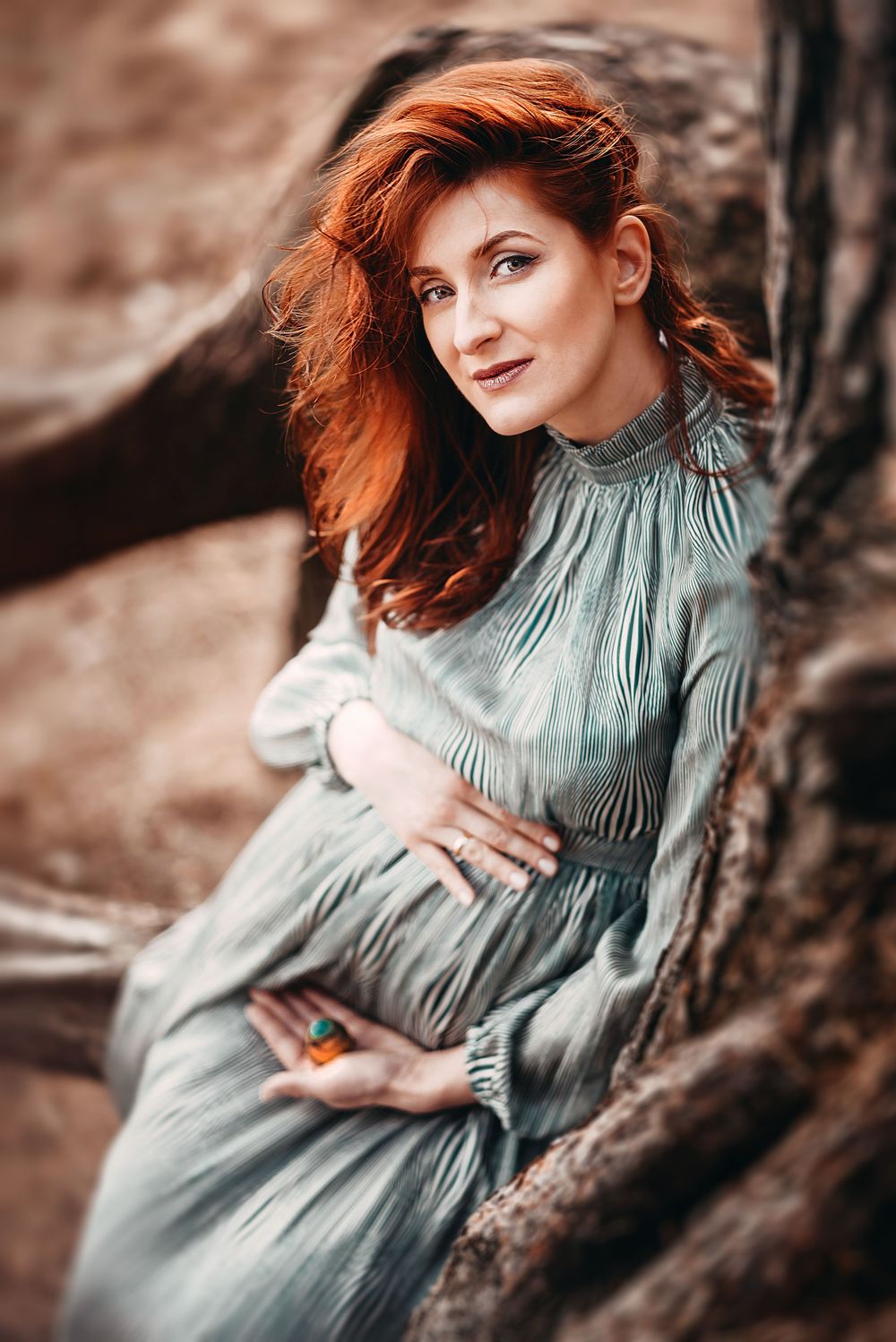 Pregnant redhead woman in teal dress sitting against tree roots, cradling belly outdoors.