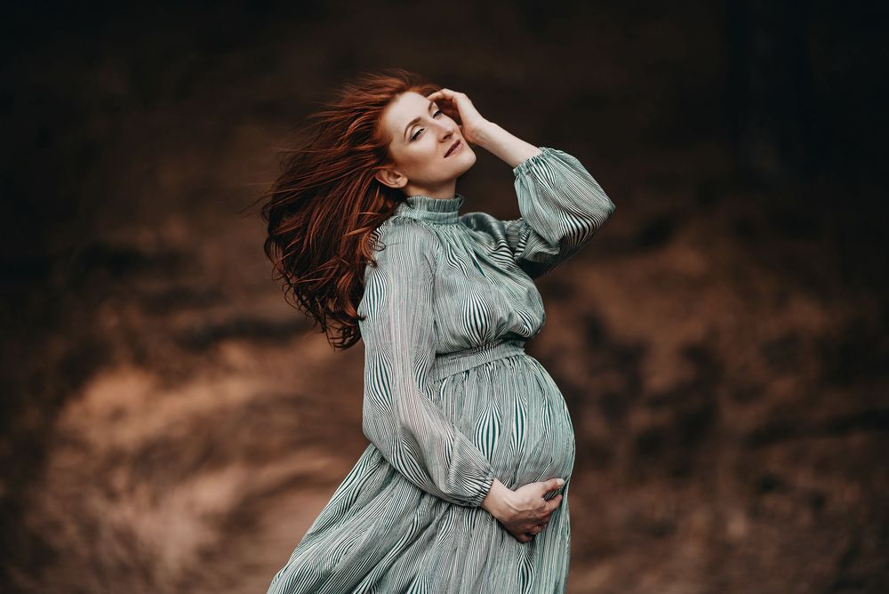 Pregnant redhead woman in teal dress poses gracefully outdoors with flowing hair in a blurred natural background.