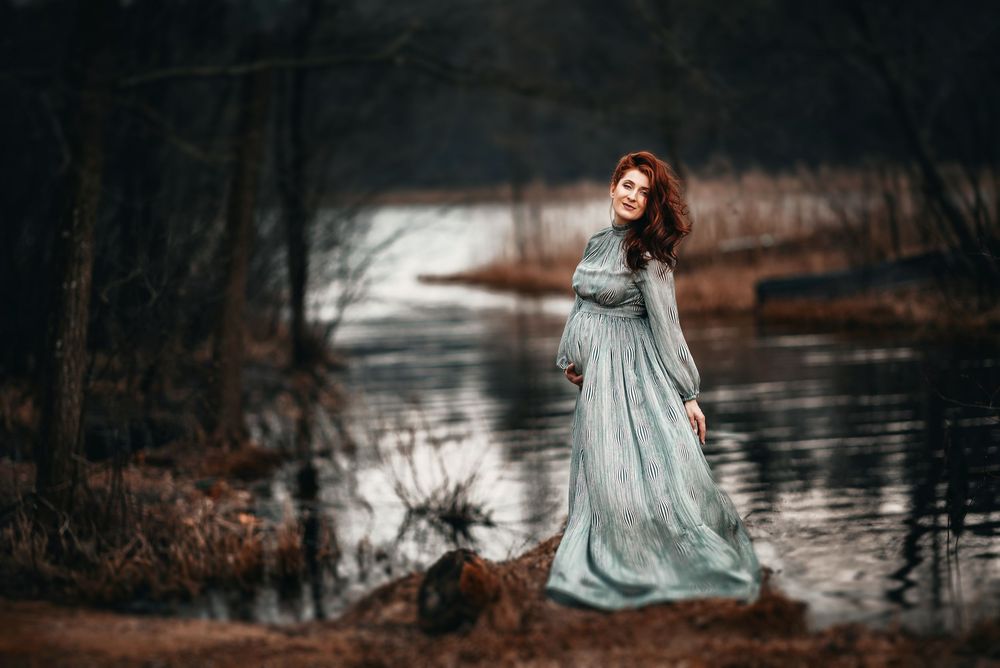 Pregnant woman in teal gown poses gracefully by a tranquil forest lake during an outdoor maternity photo shoot.