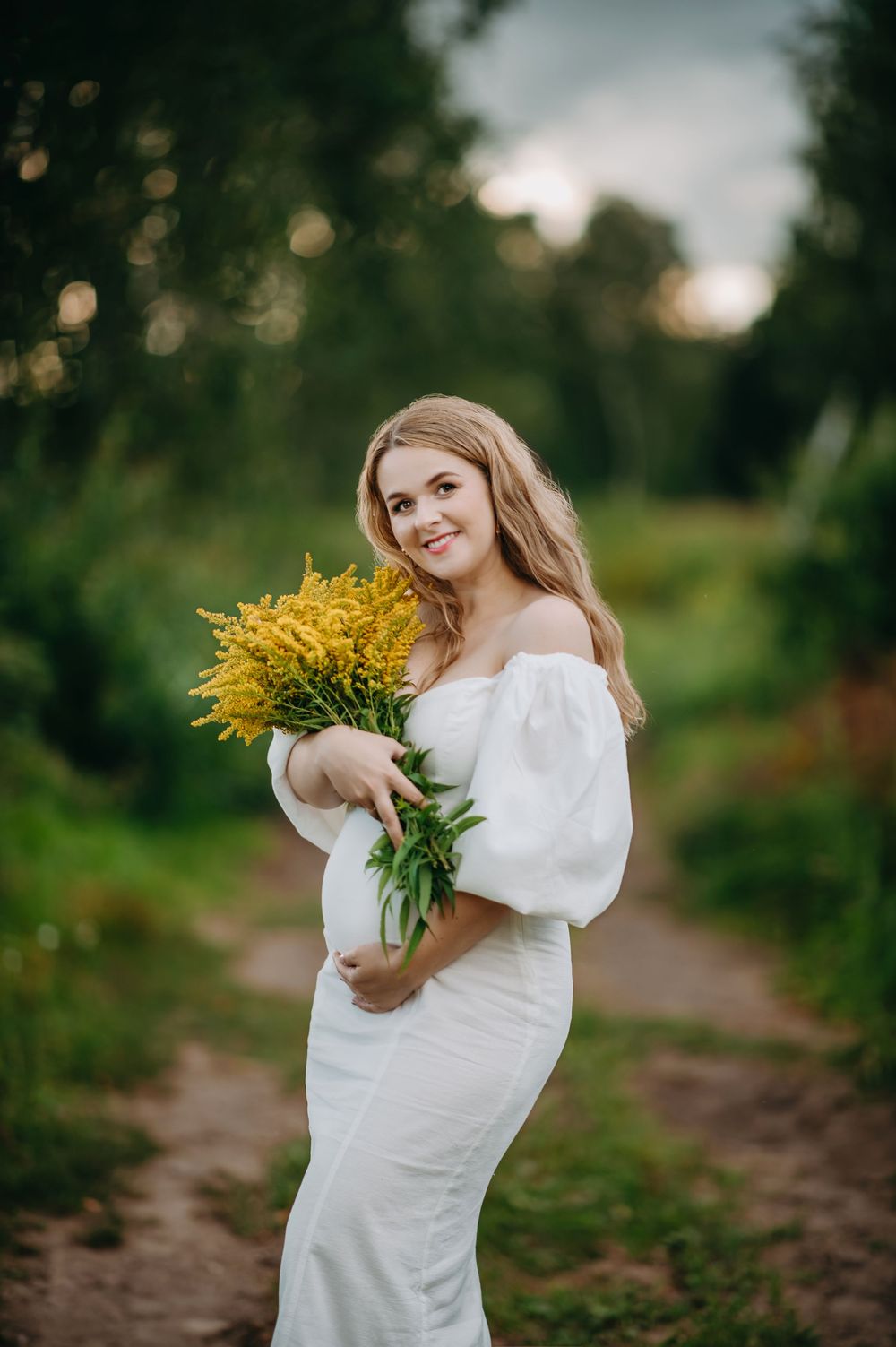 Pregnant woman in white off-shoulder dress holds yellow wildflowers on a green nature path.