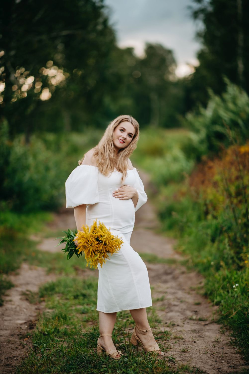 Pregnant woman in white off-shoulder dress holding yellow flowers on a scenic dirt path surrounded by lush greenery.