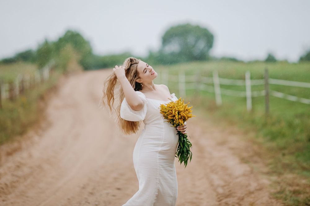 Pregnant woman in white dress holding yellow flowers on a rural dirt road.
