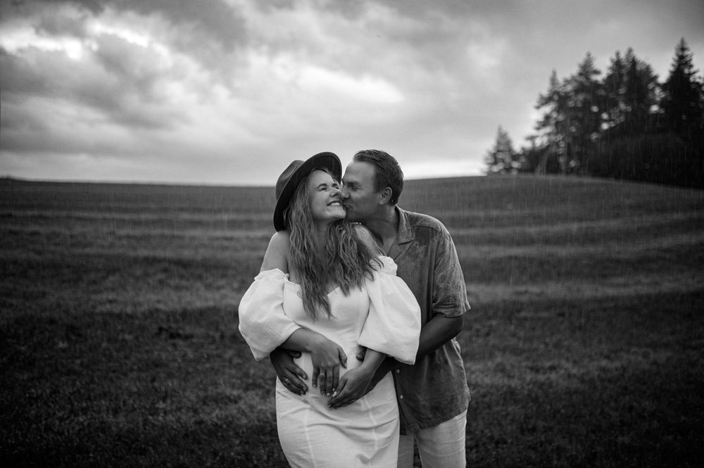 Black and white photo of a couple embracing in a field, man kissing womans cheek under dramatic cloudy sky.