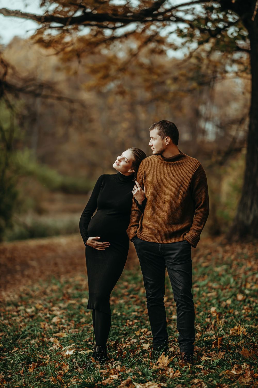 Pregnant couple embracing in autumn park, woman in black dress holding belly, man in brown sweater among fallen leaves.