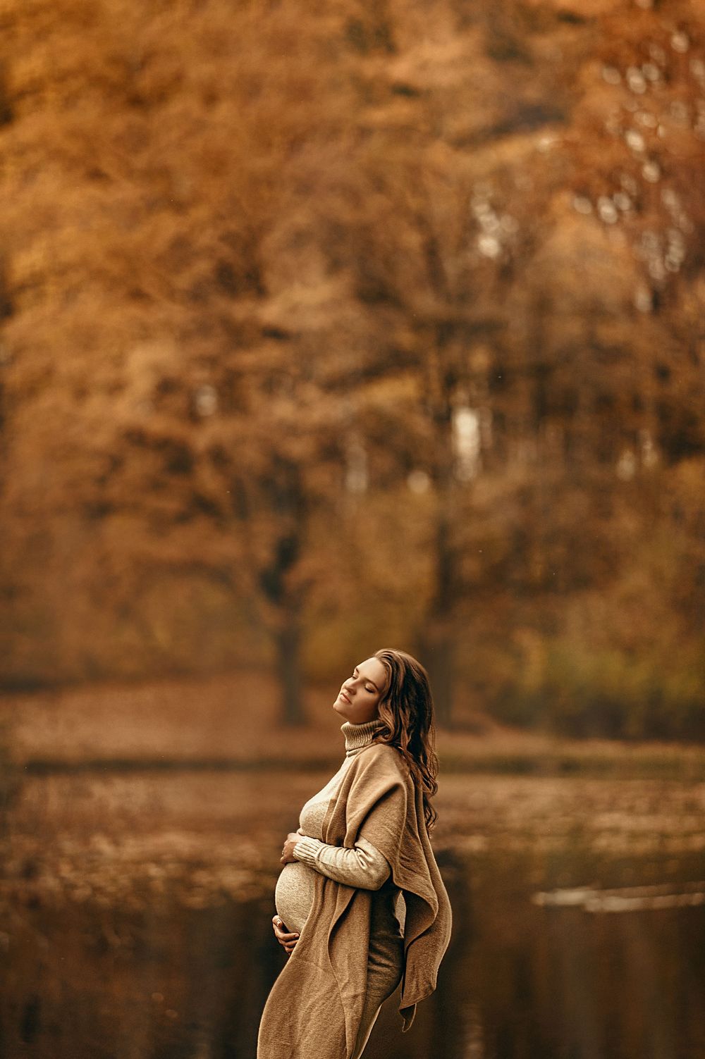 Pregnant woman in beige wrap coat cradles her belly outdoors among golden autumn trees near a tranquil lake.