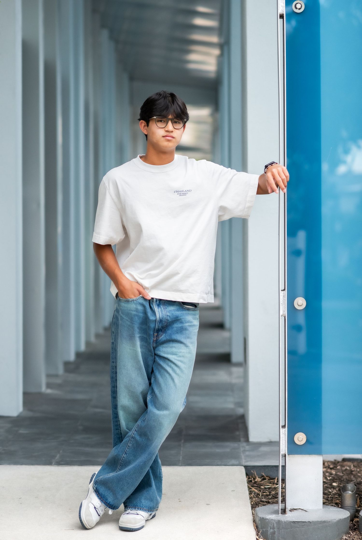 Young man in white tee and wide-leg jeans leaning against blue pillar in modern corridor.