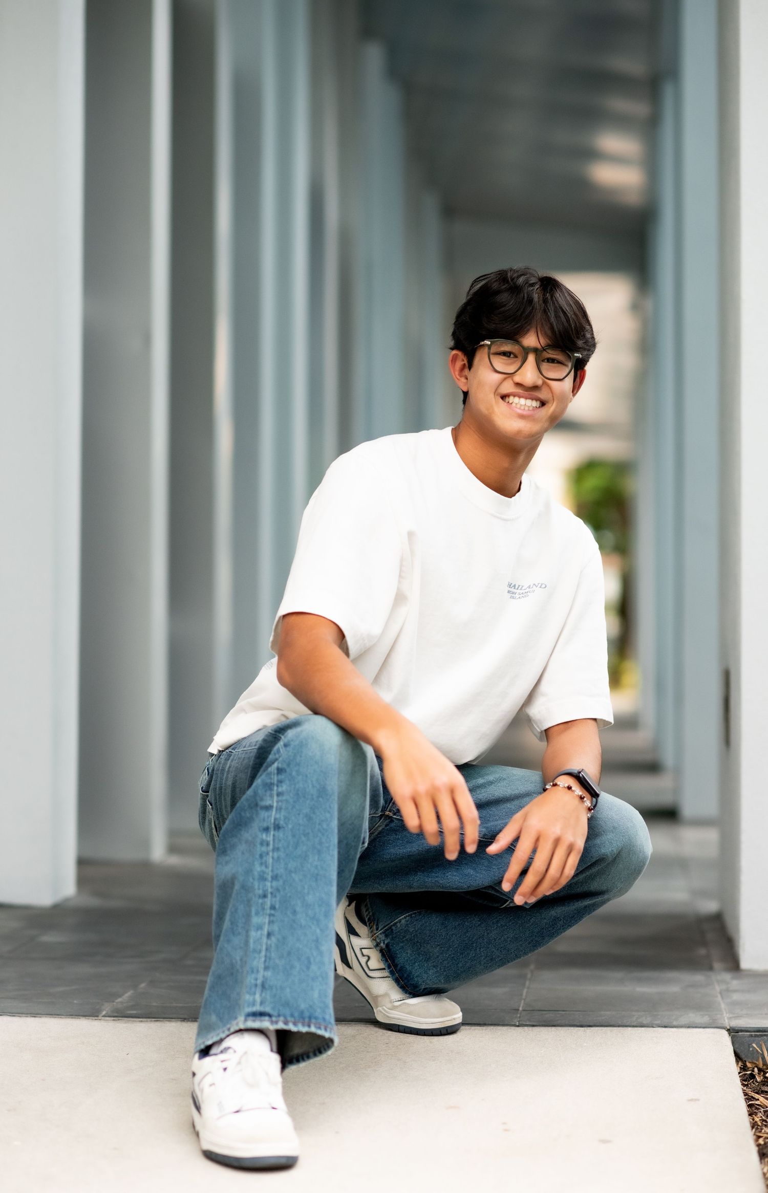 Smiling young man in white t-shirt and jeans crouching in a modern hallway corridor.