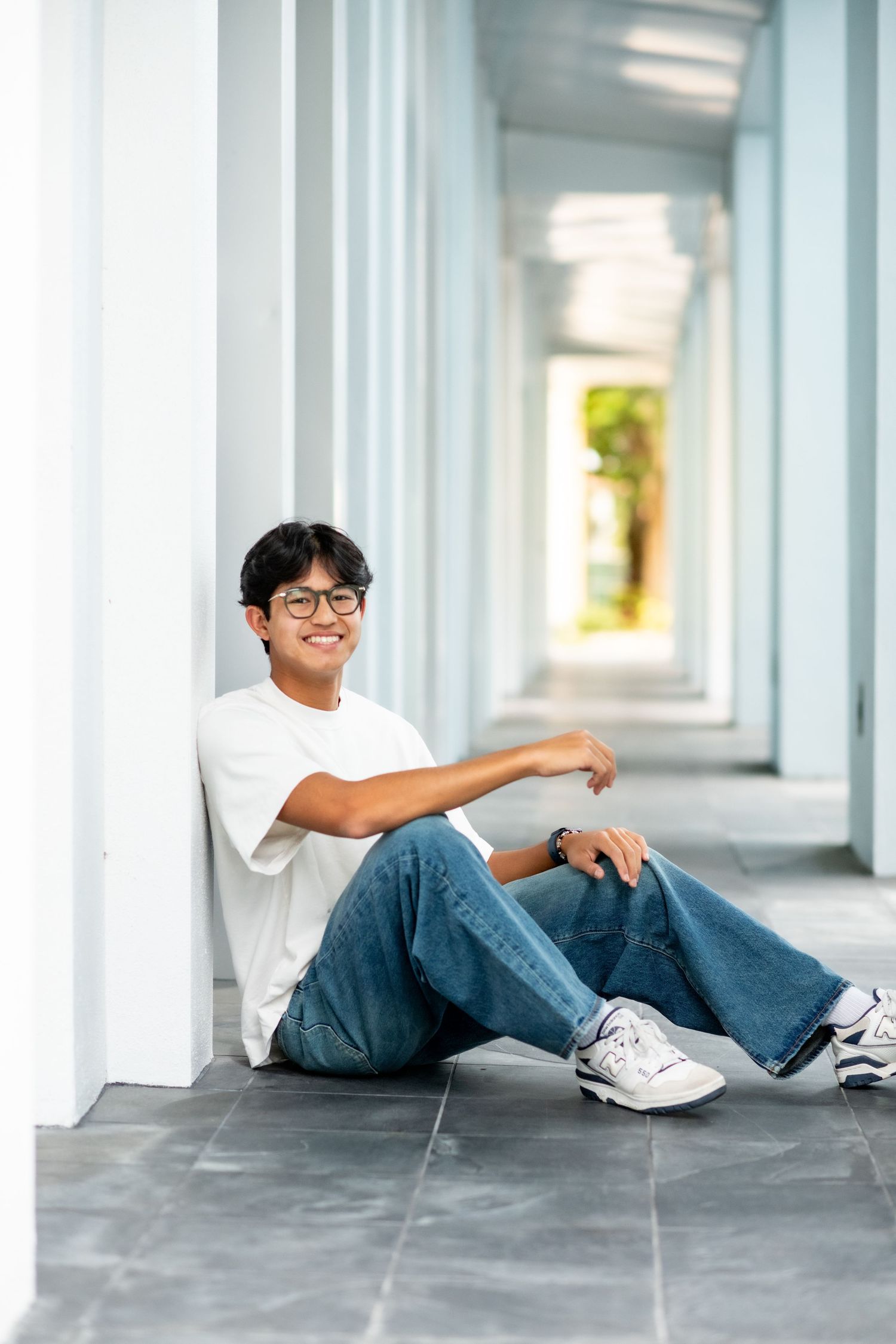 Smiling teen in white tee and jeans sitting in a bright corridor.