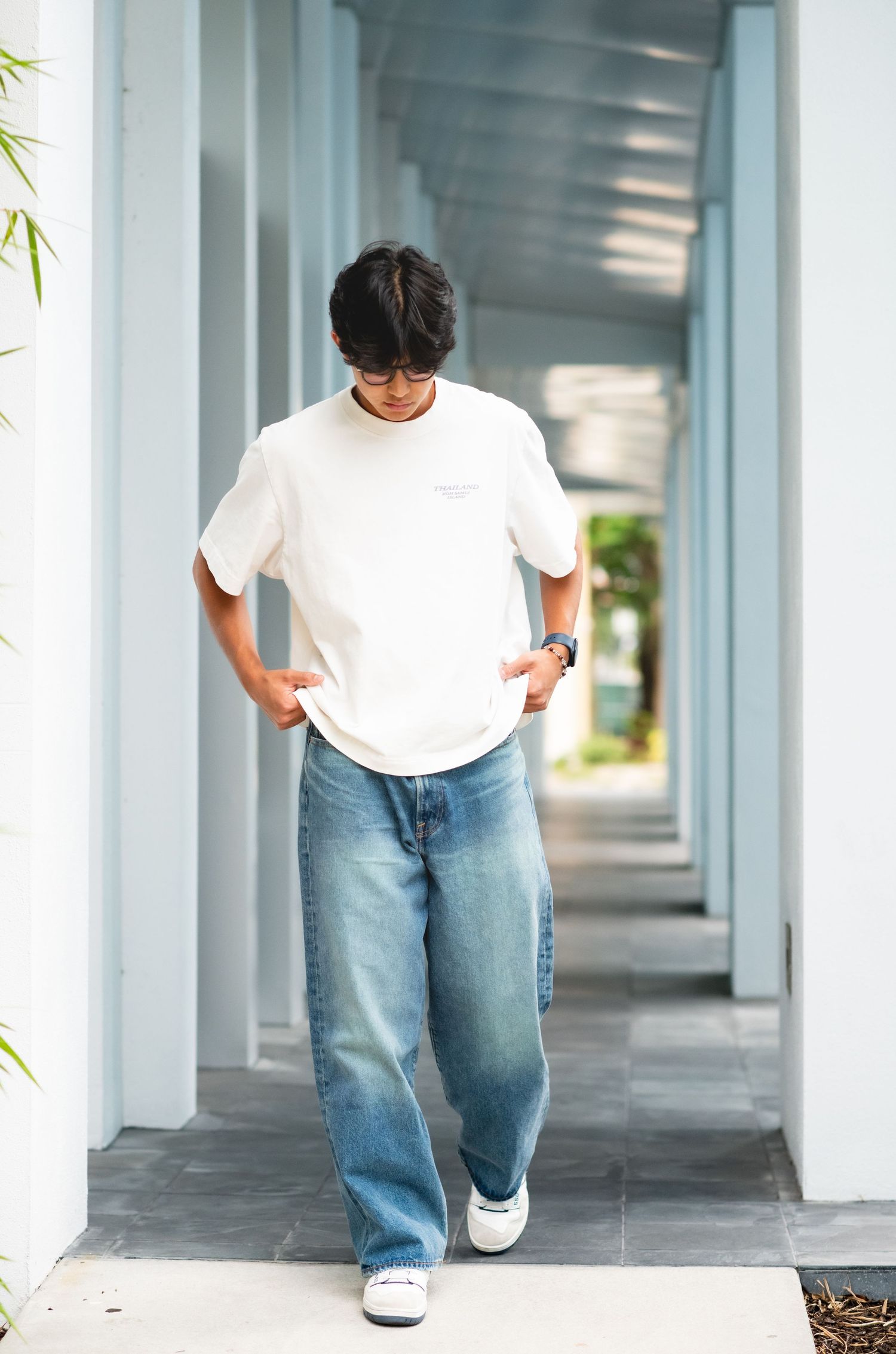 Young man in white oversized tee, wide-leg jeans, and sneakers walking through a bright corridor.
