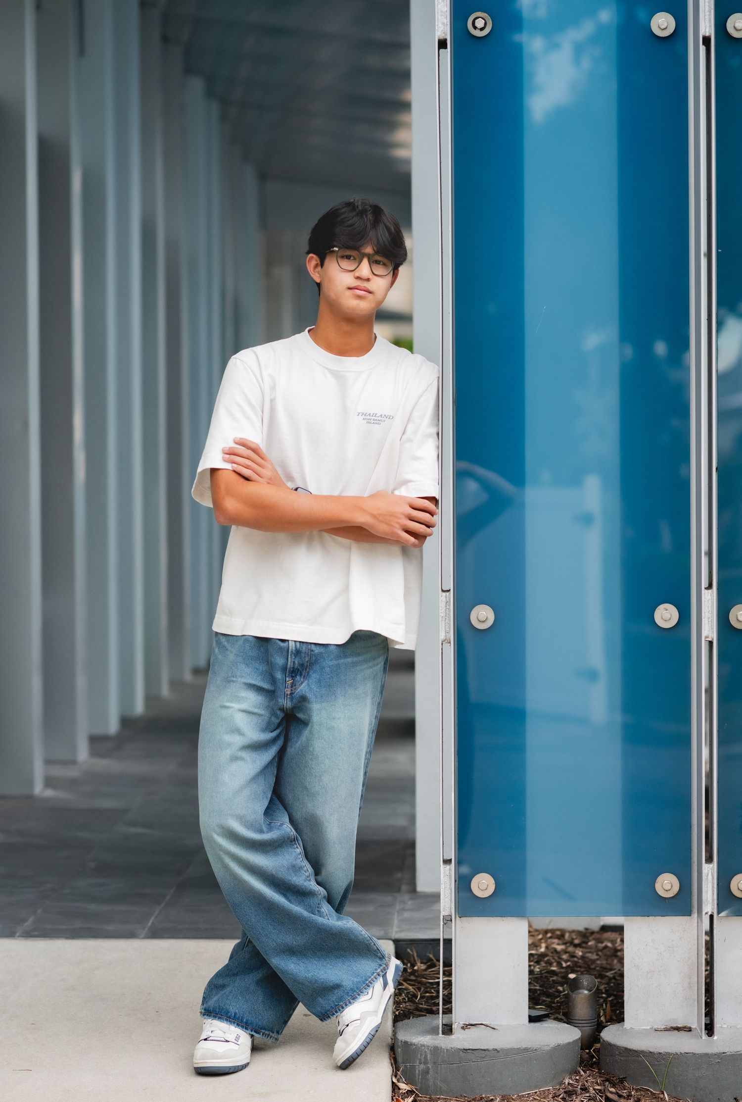 Teen boy in white t-shirt and jeans standing with arms crossed by blue column outdoors.