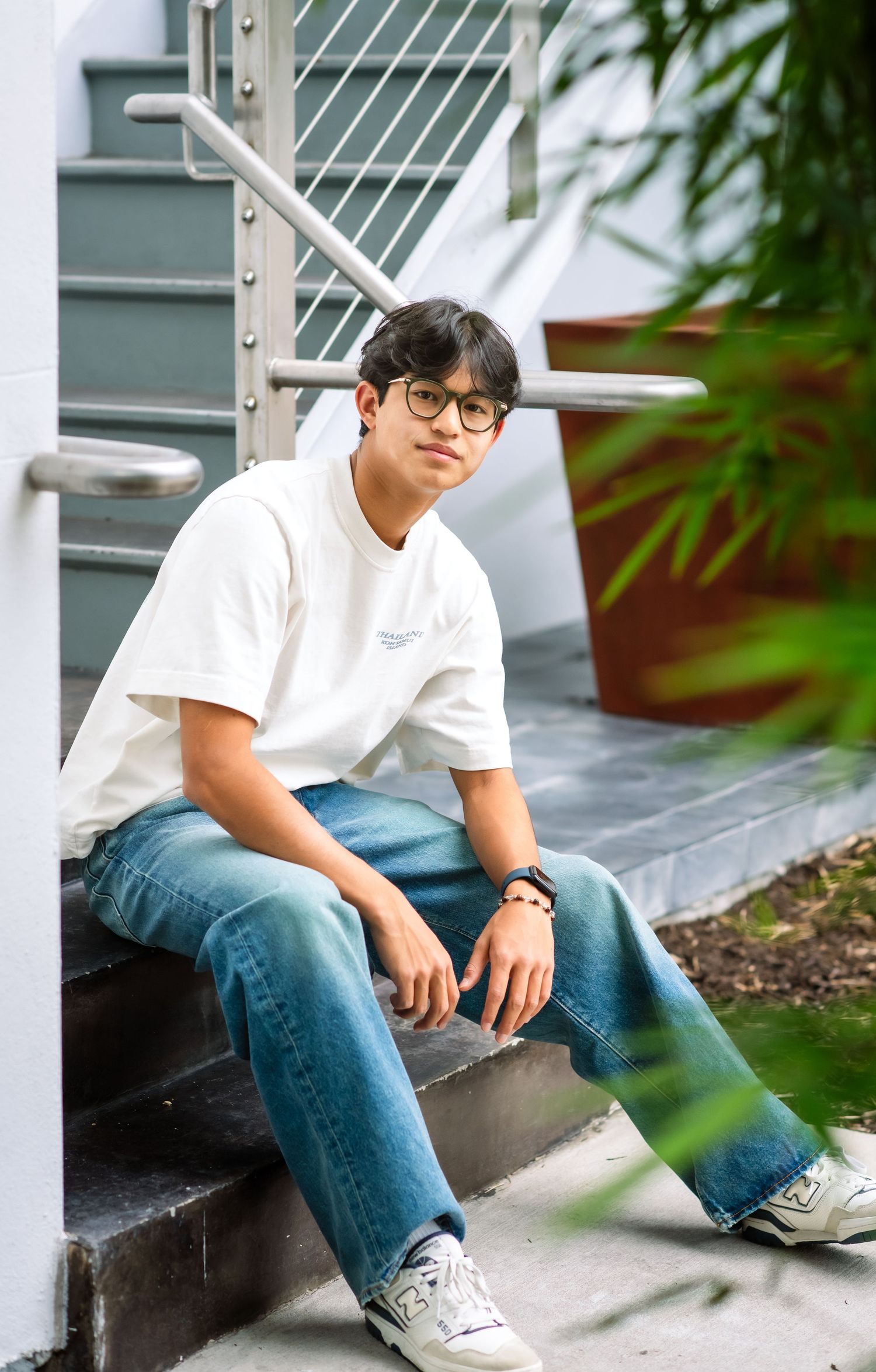 Young Asian man in white tee and jeans sitting on outdoor stairs.