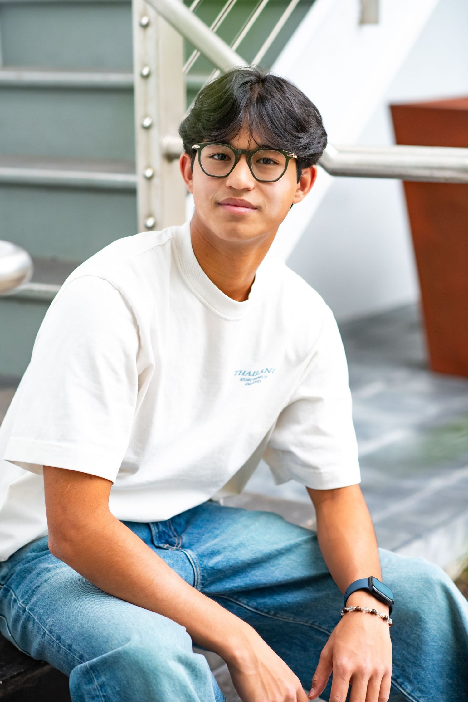 Young man in white t-shirt and jeans with glasses sits on steps, smiling casually outdoors.