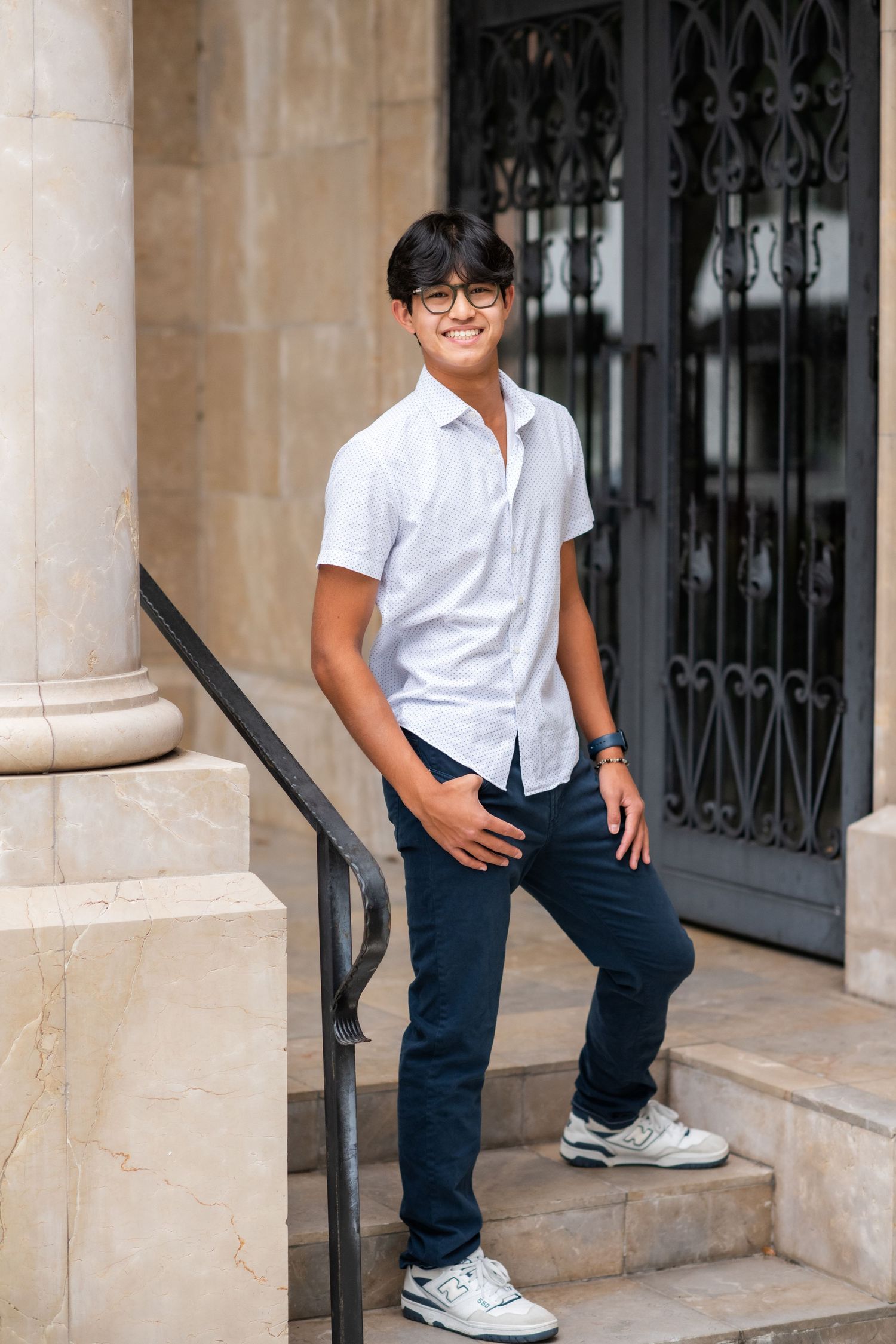 Smiling teen in white shirt and navy pants posing on stone steps by ornate door.