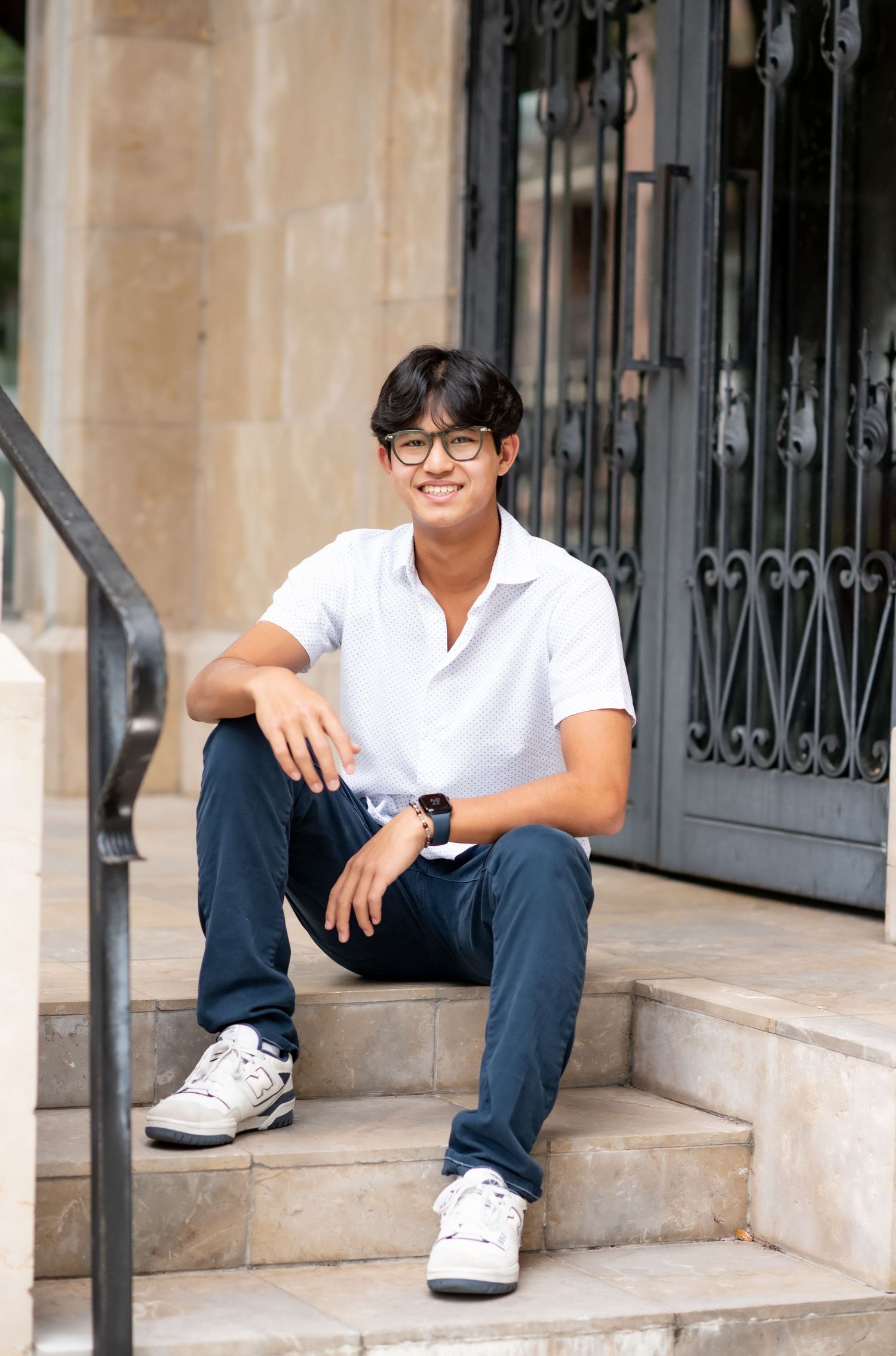 Smiling young man in white polo and navy pants sitting casually on stone steps outdoors.
