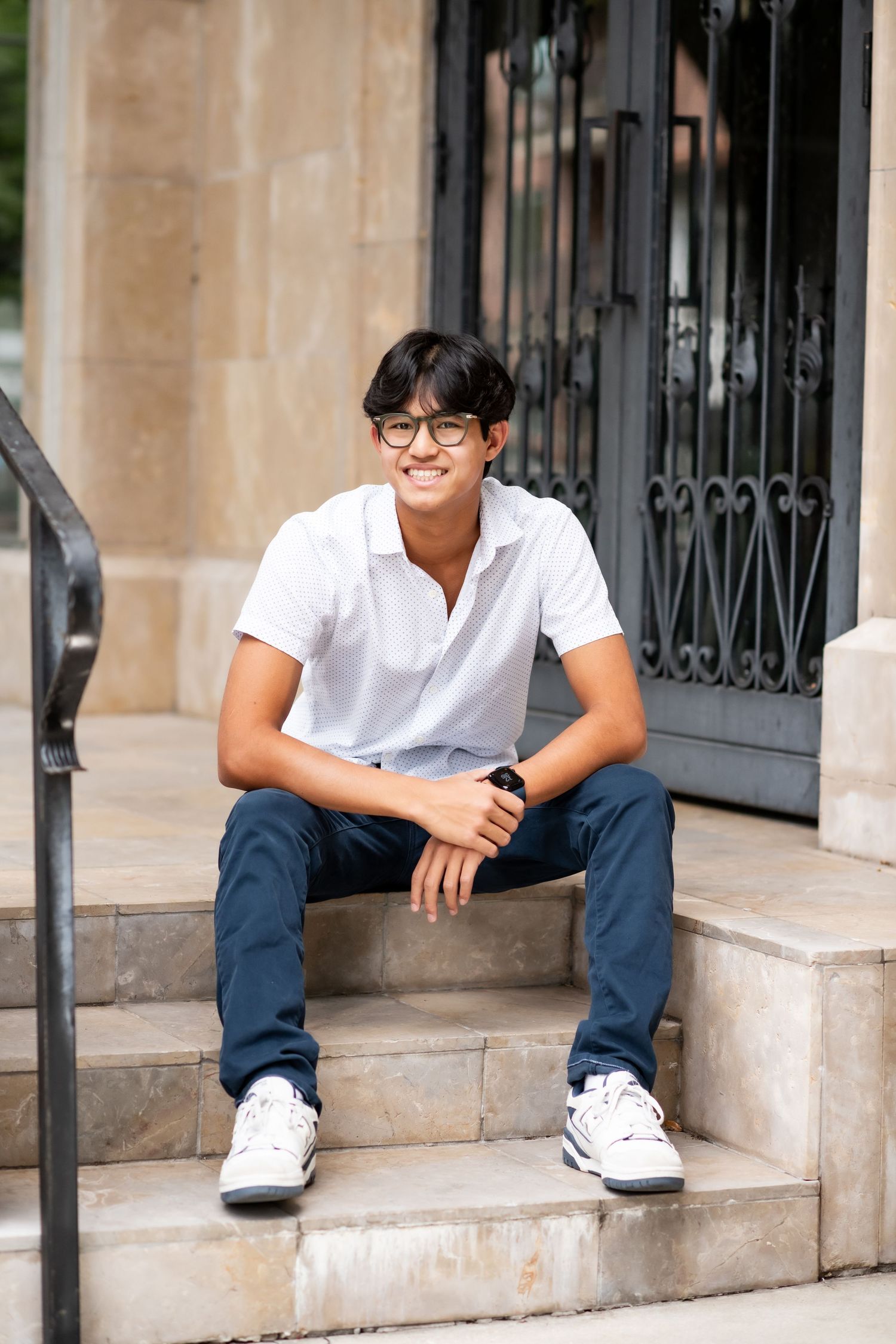 Young man in white polo shirt and navy pants smiling while seated on stone steps outside.