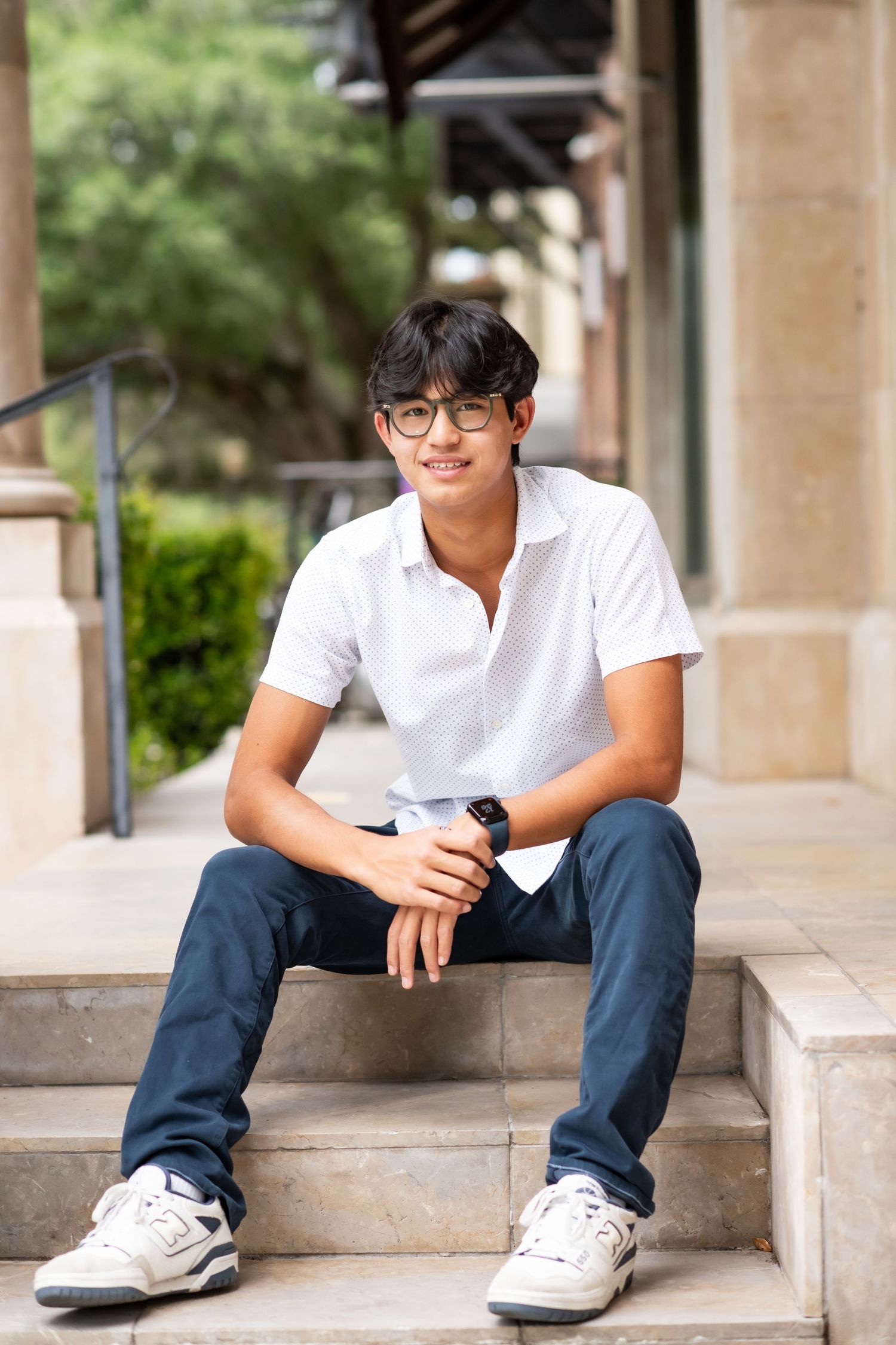 Smiling teenage boy in white polo shirt and navy pants sitting on stone steps outdoors.