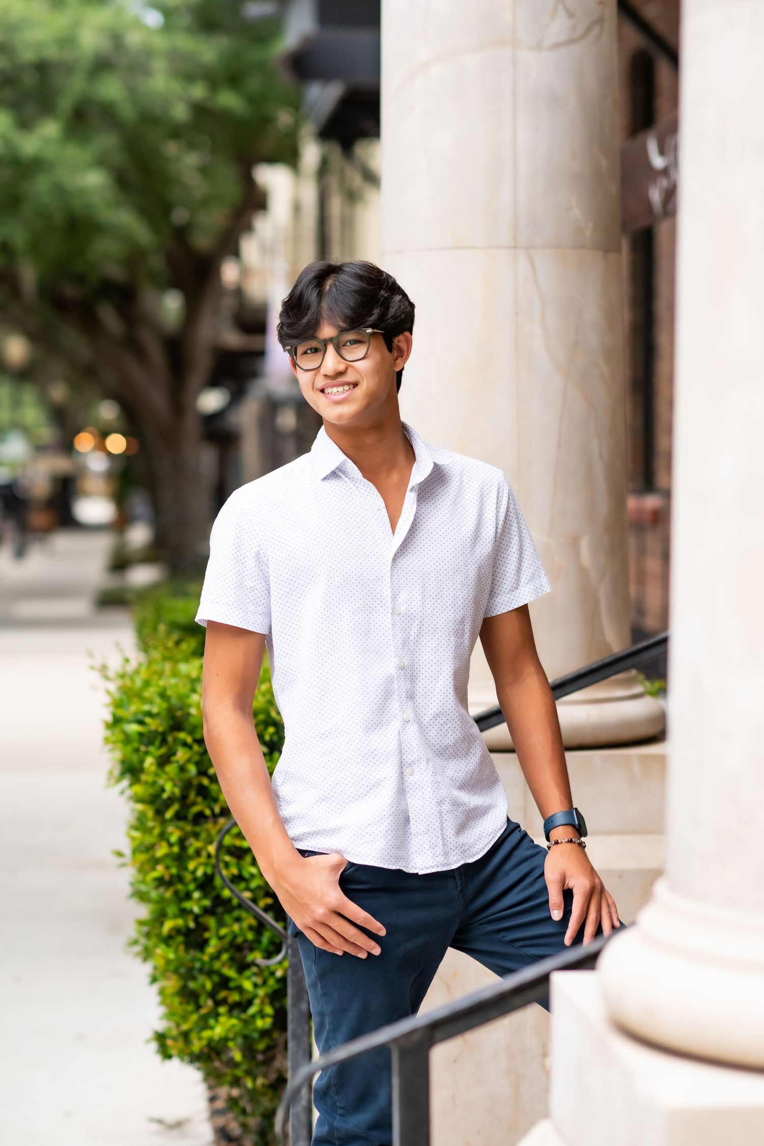 Smiling young Asian man in white short-sleeve shirt and glasses leaning on railing outdoors.