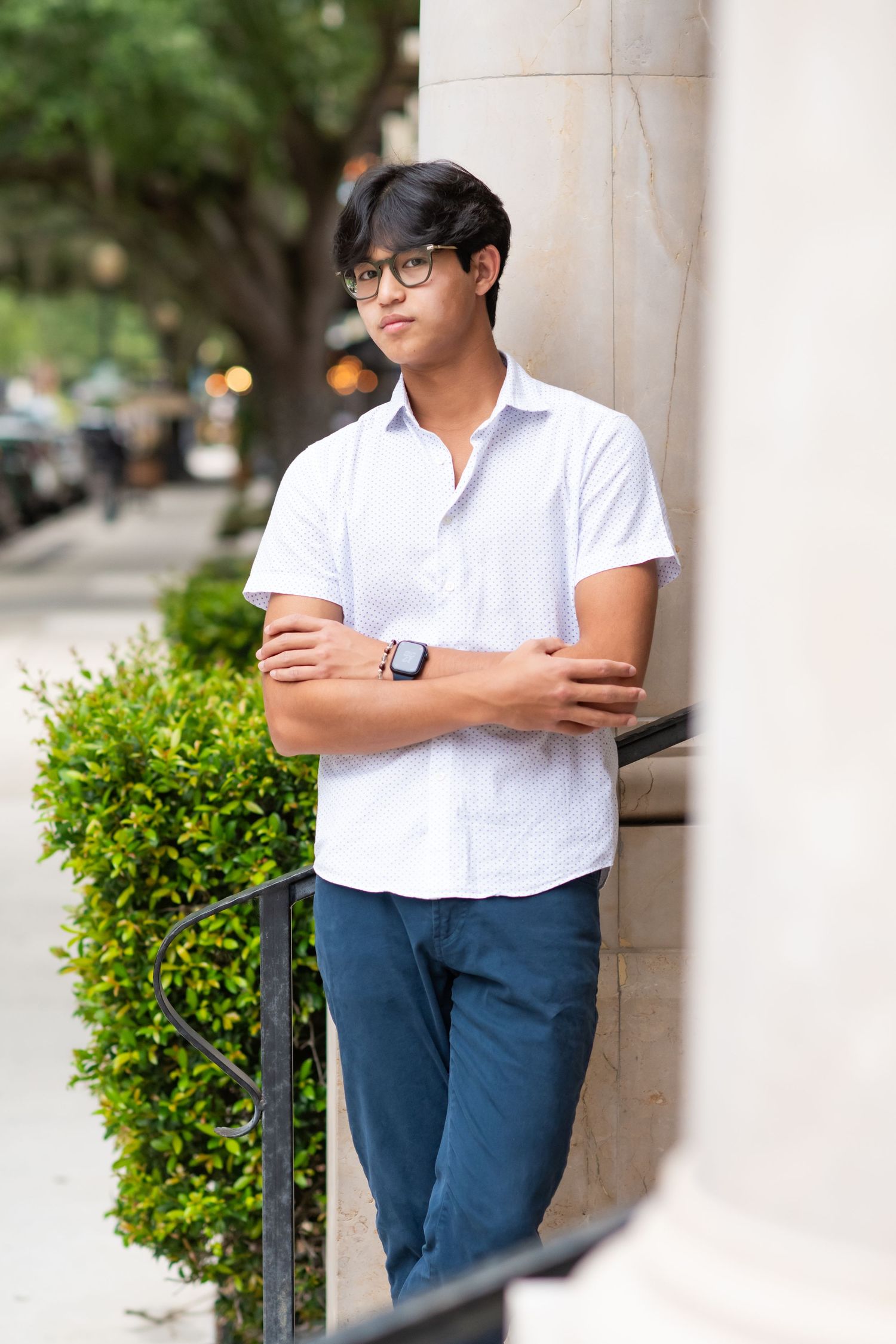 Teen boy in white shirt with smartwatch leaning against column with arms crossed.