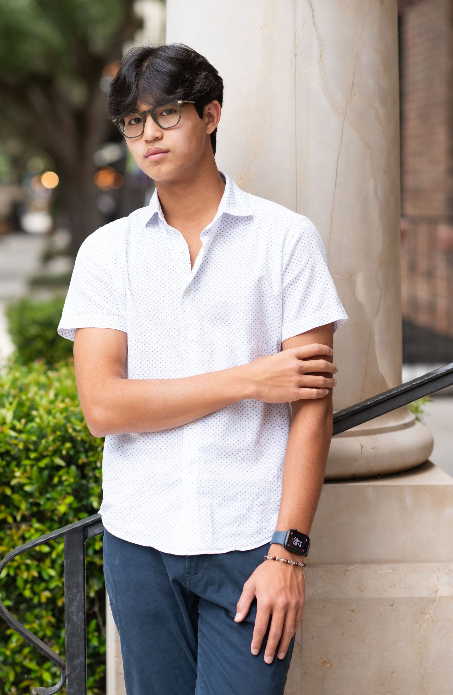 Young man in white short-sleeve shirt and glasses leaning against a column outdoors.