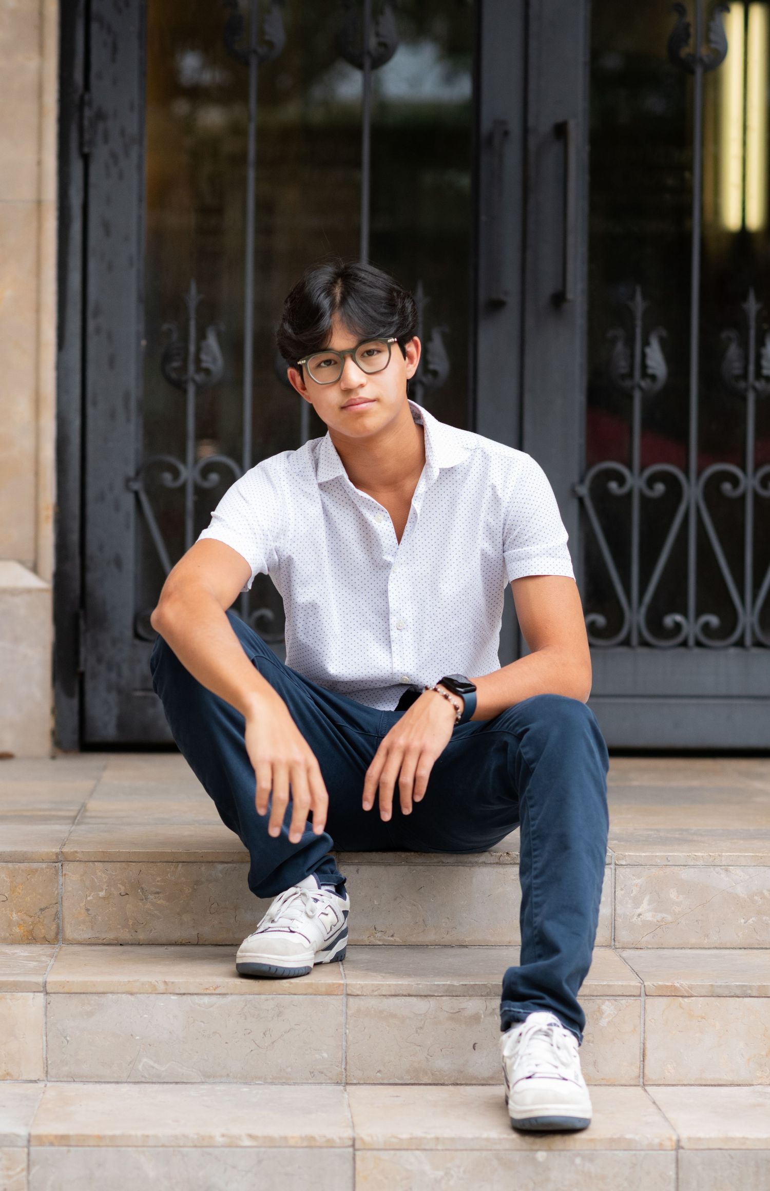 Teen boy in white polo sitting on stone steps by ornate doors.