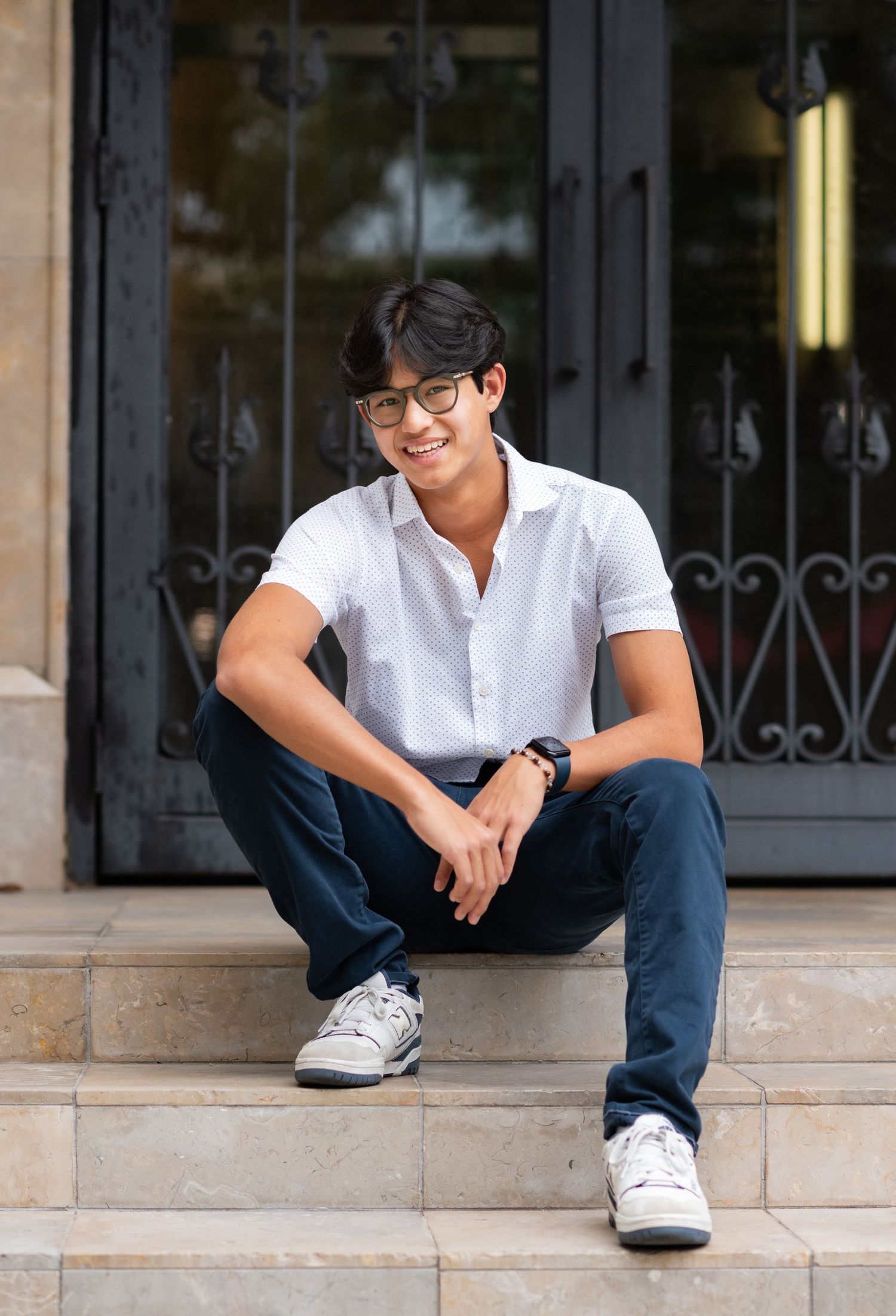 Smiling young man in white shirt and jeans sitting on stone steps outdoors.