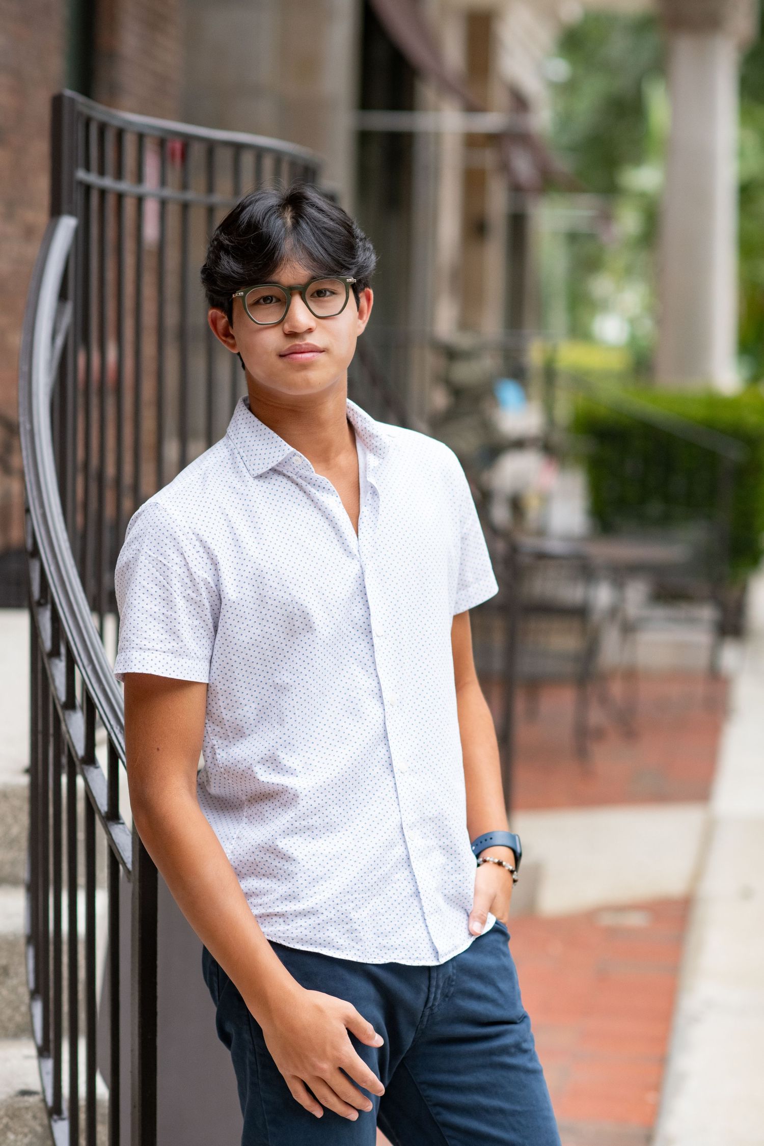 Teen boy in white button-up shirt leans against iron railing on urban sidewalk.