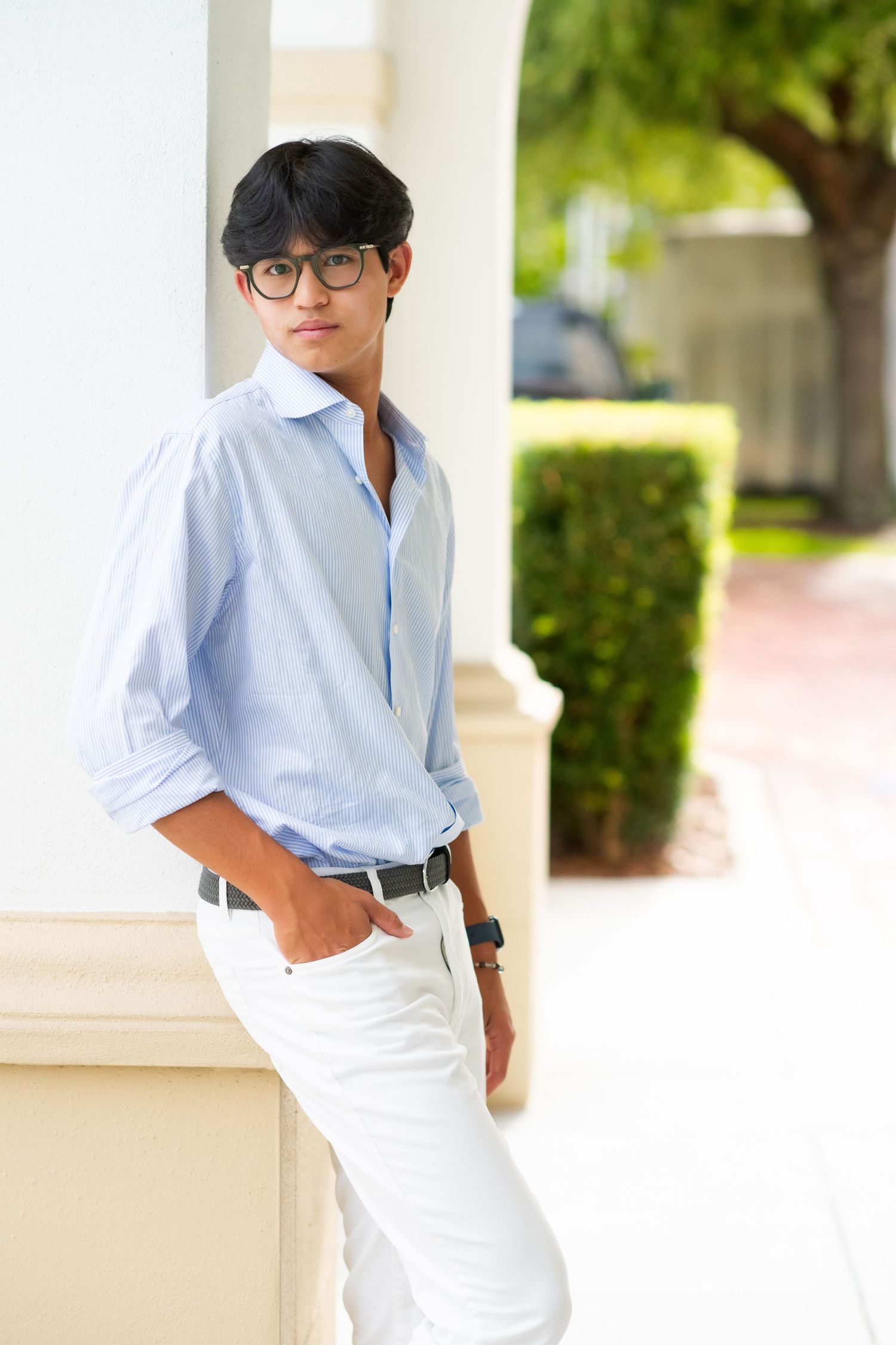Stylish young man in light blue shirt and white pants leaning against a column outdoors.