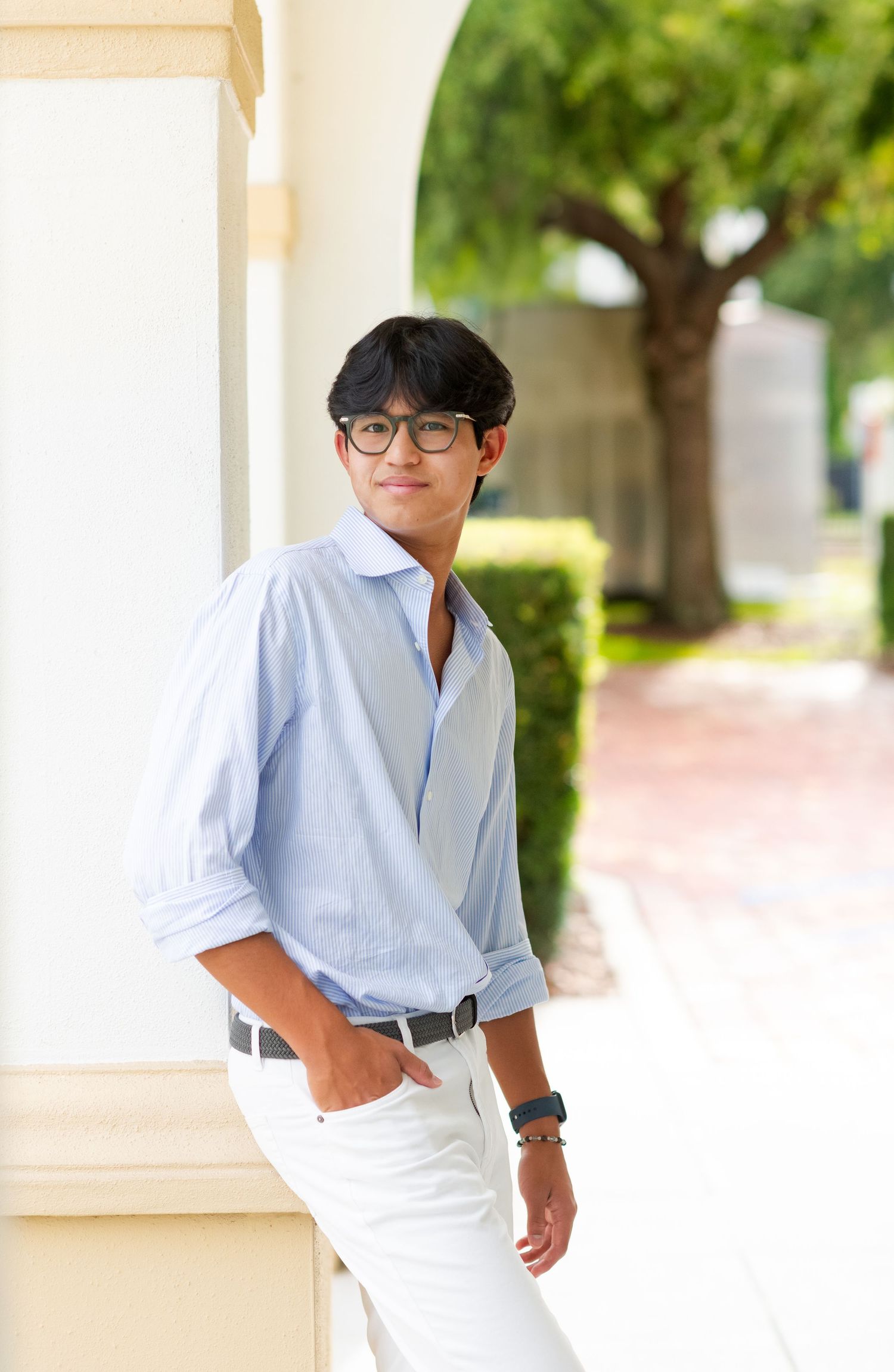 Young man in light blue button-up shirt and white pants leaning against a column outdoors.