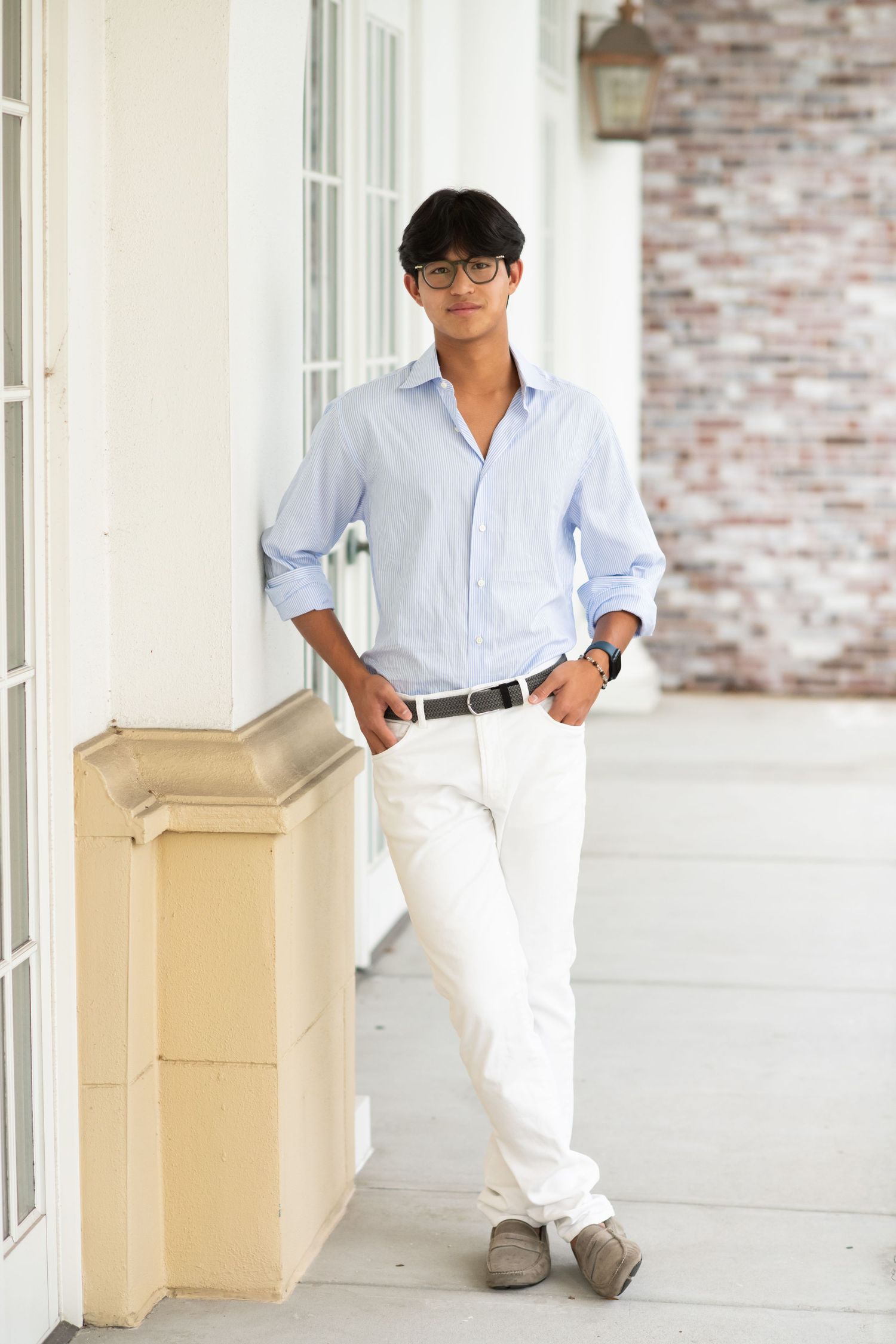 Young man in light blue dress shirt and white pants leans against column on porch.