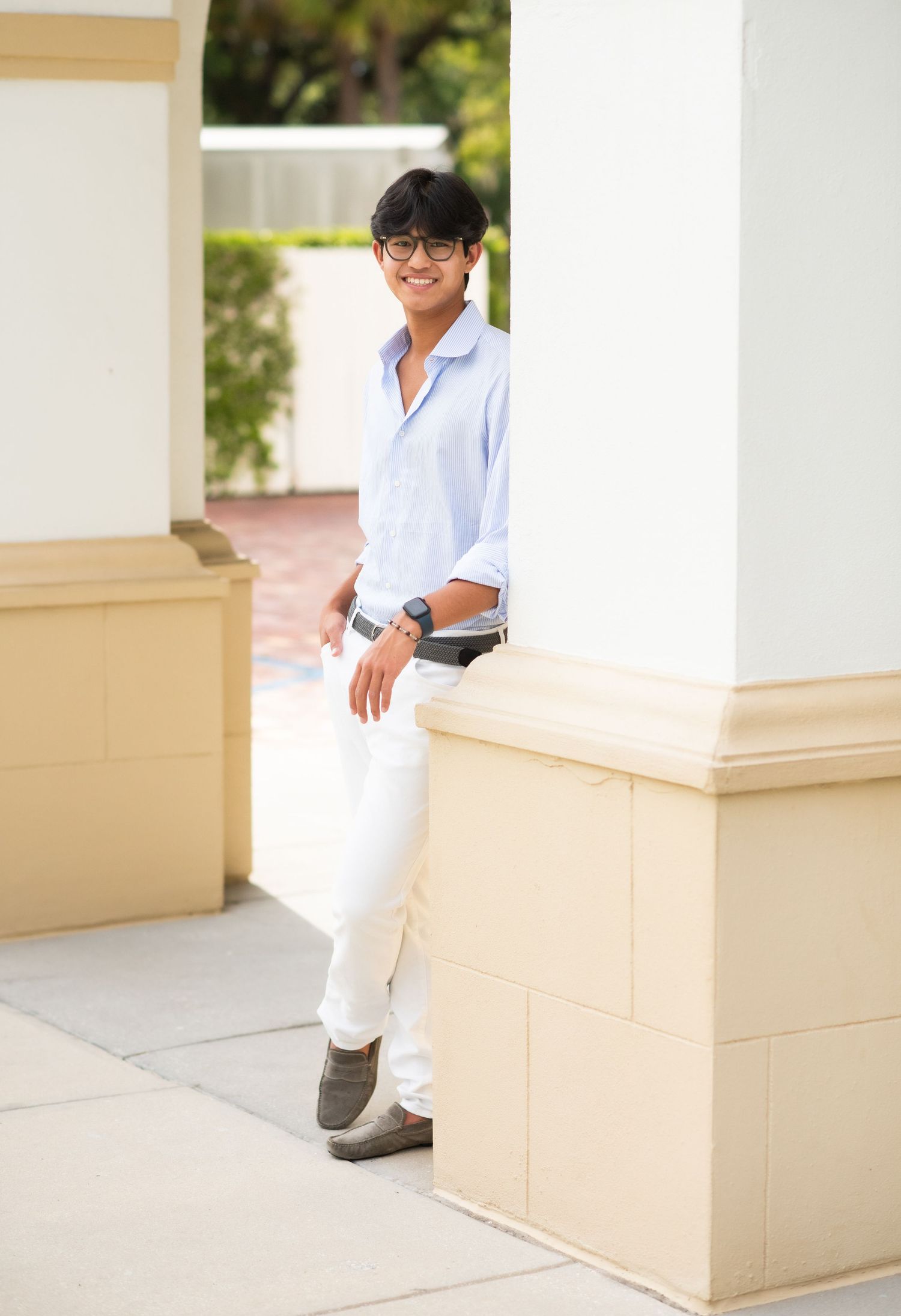 Young man in light blue shirt and white pants leaning against a column outdoors, smiling confidently.