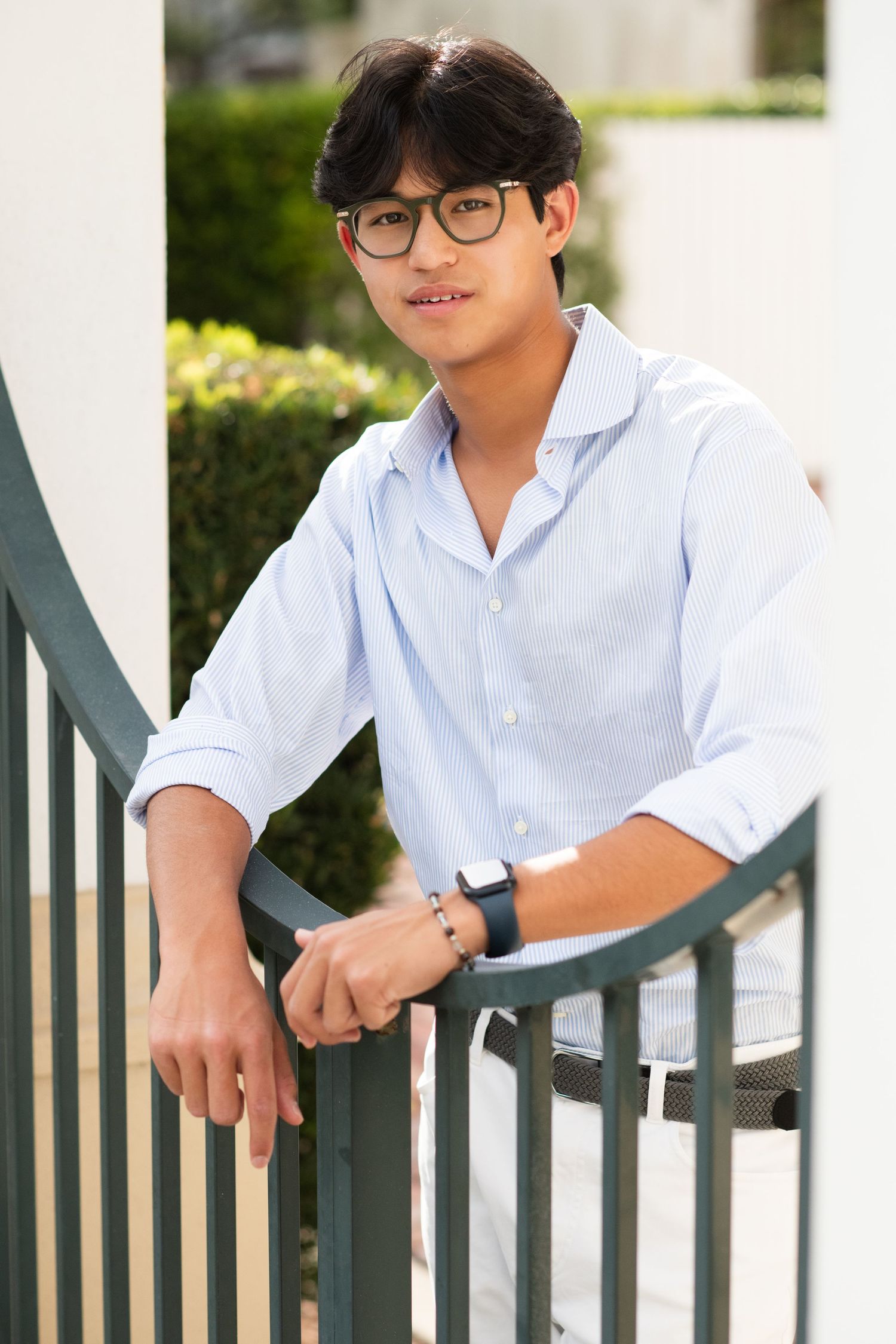 Smiling young Asian man in light blue shirt leaning on green railing outdoors.