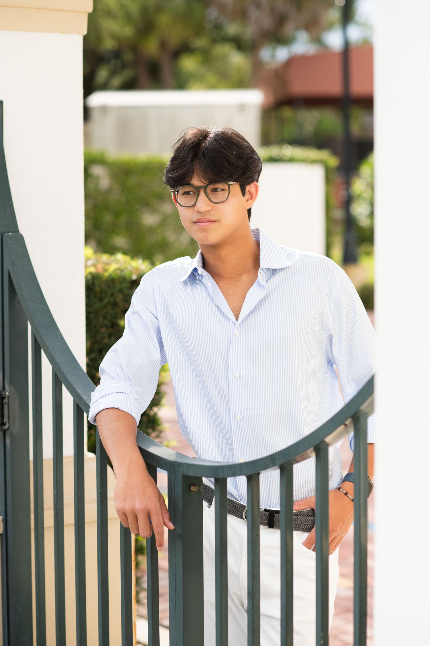Young man wearing glasses in a light blue shirt leaning on a green gate in an outdoor setting.