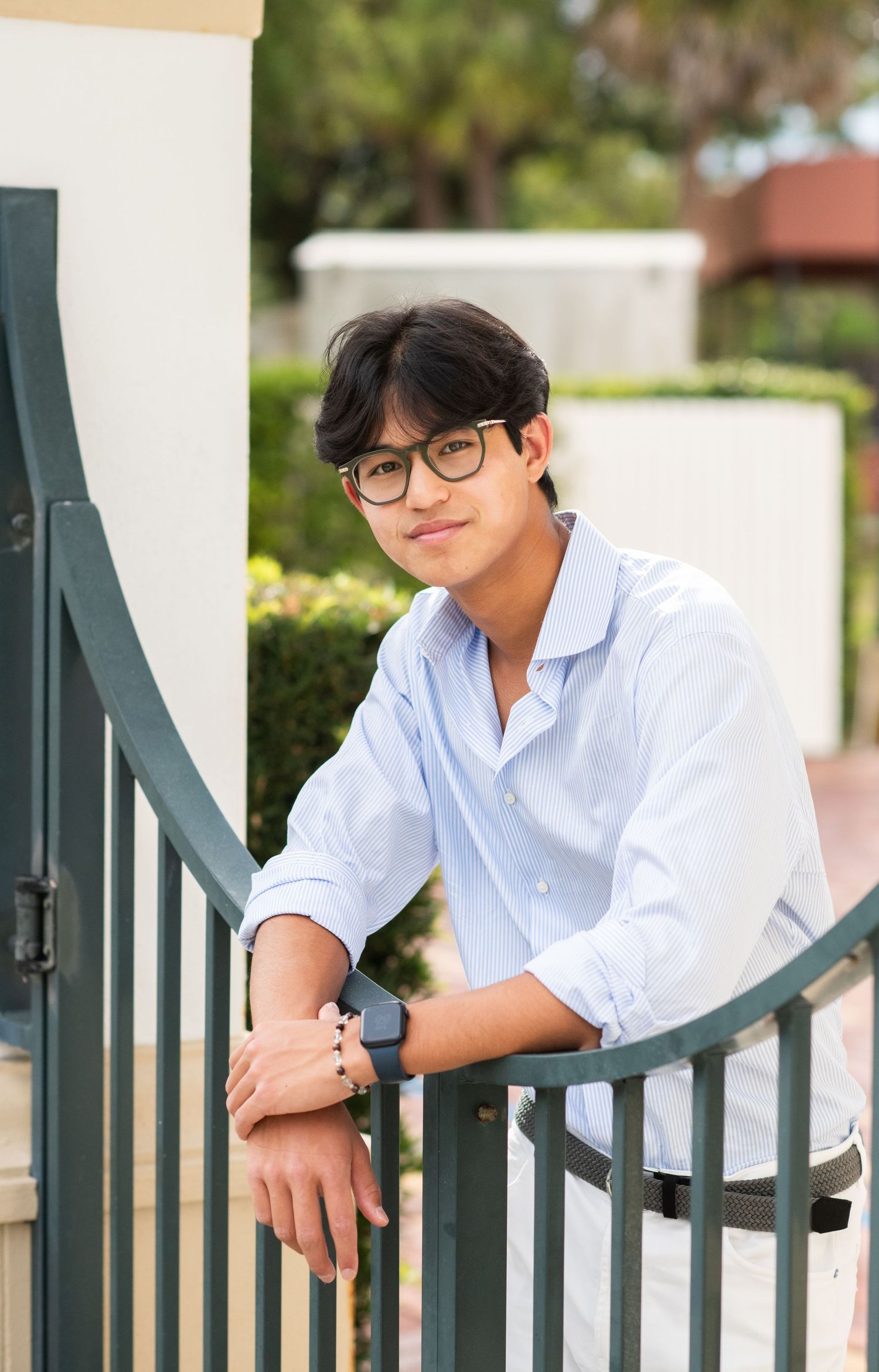 Young Asian man in glasses and white shirt leaning on a green railing, smiling outdoors.