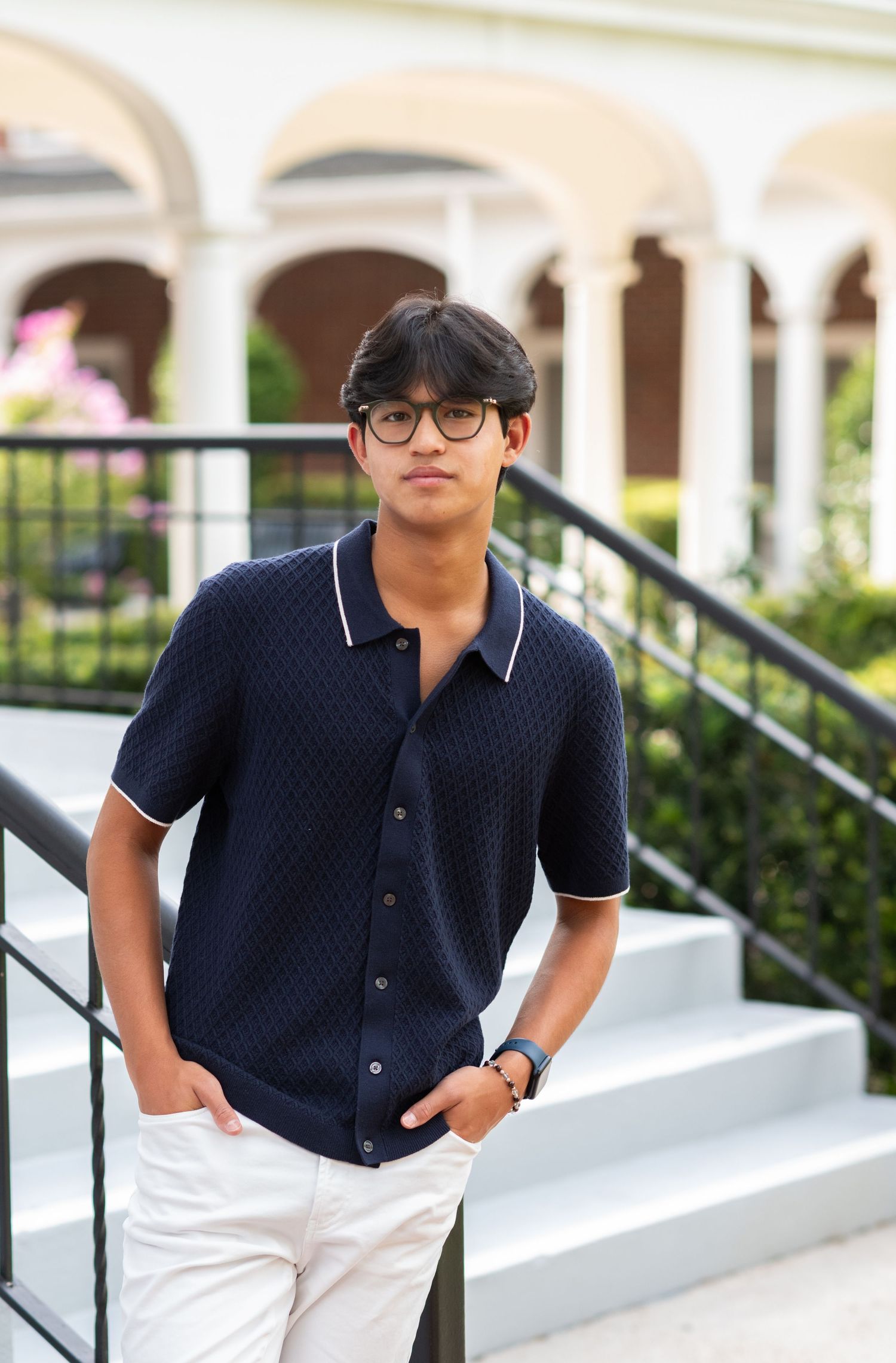 Young man in navy blue textured polo shirt and white pants posing on outdoor stairs.