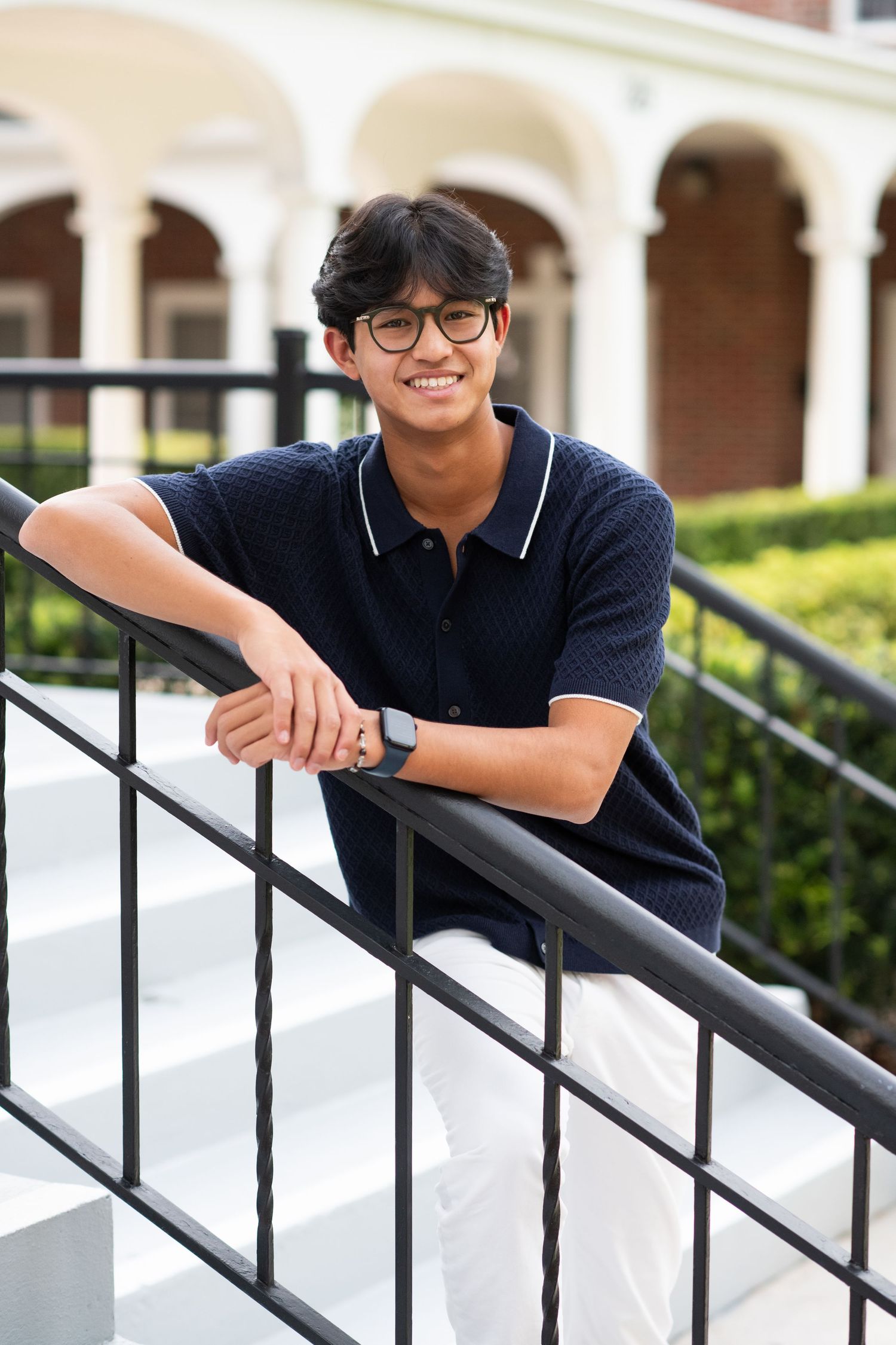 Smiling teen boy in navy polo leans on black railing on outdoor brick steps.