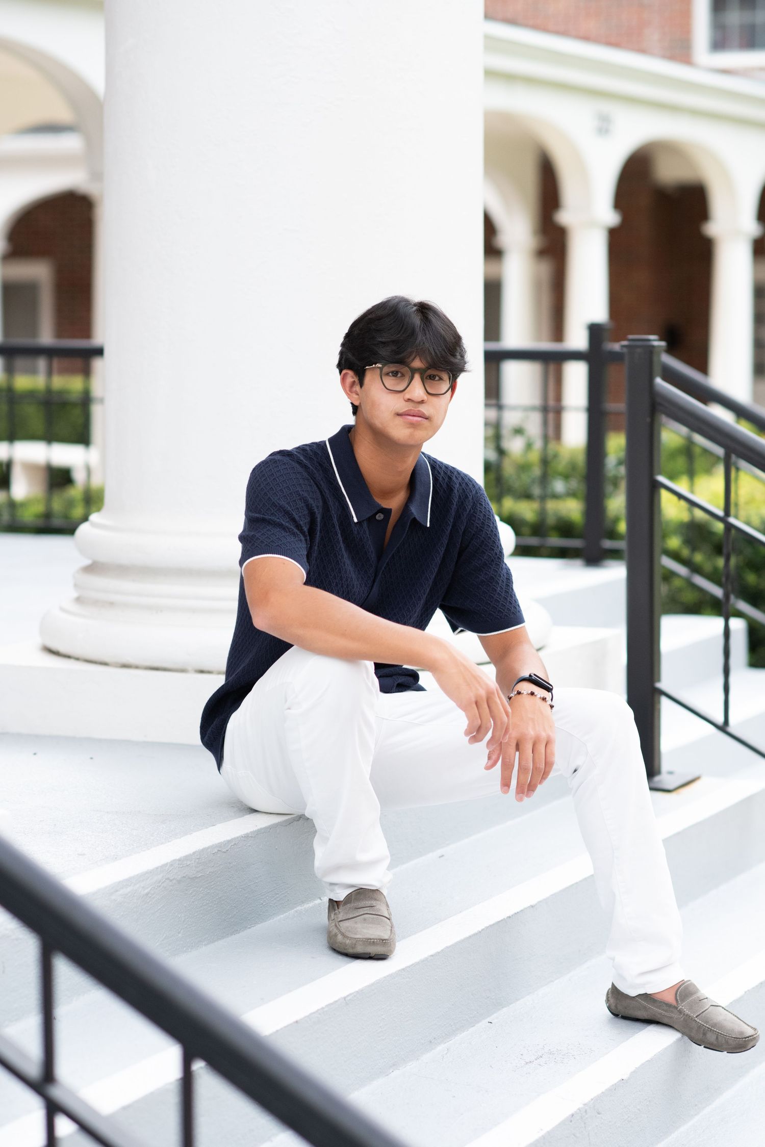 Teen boy in navy polo and white pants sits on white outdoor steps by column.