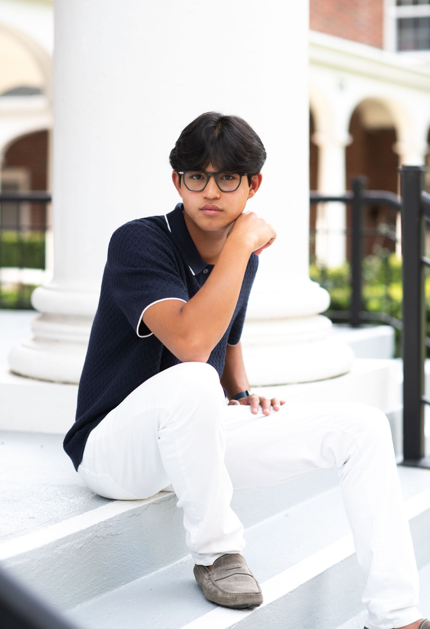 Teen in navy polo and white pants posing pensively on white steps.
