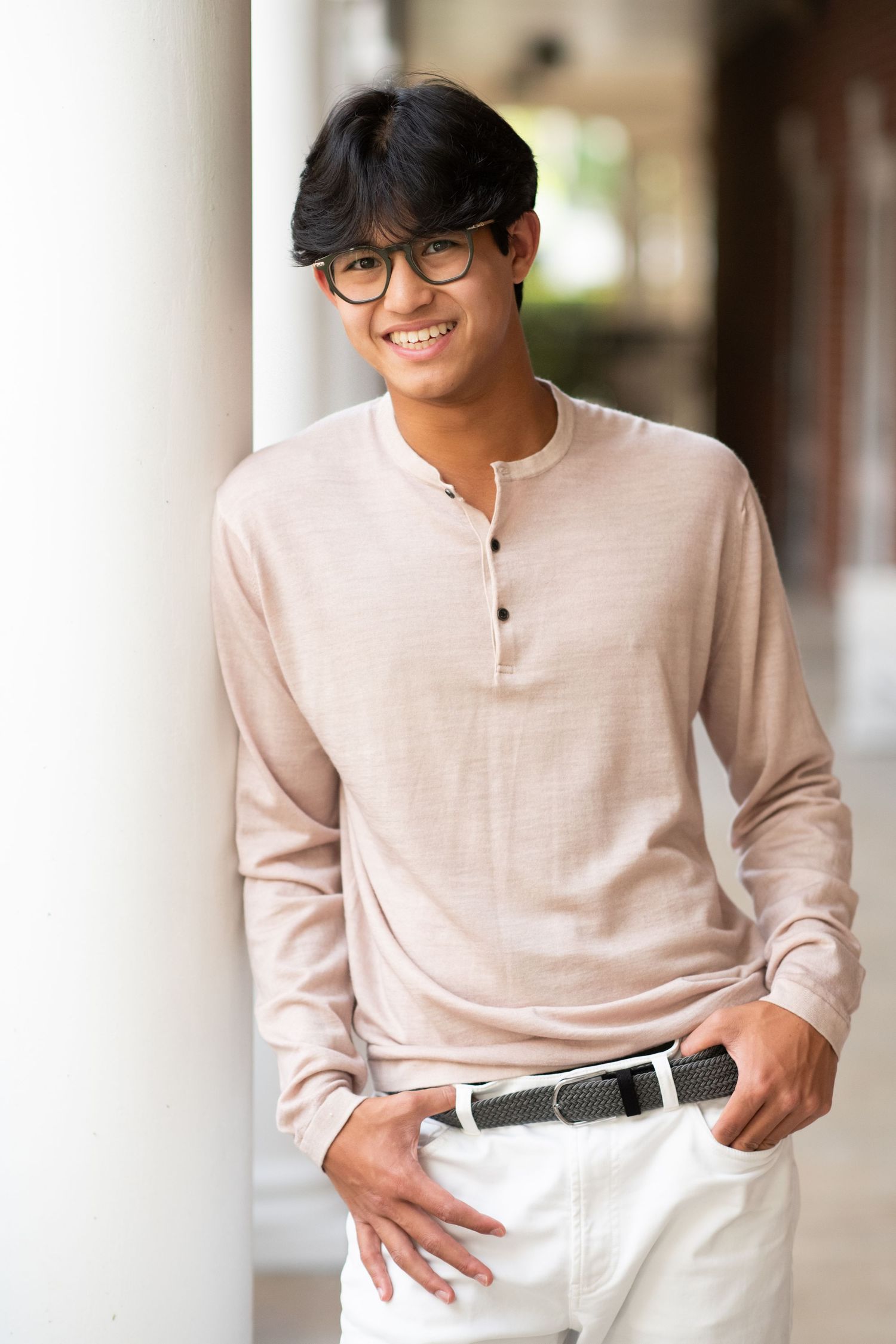 Smiling young man in beige henley shirt and white pants leaning against a white column.