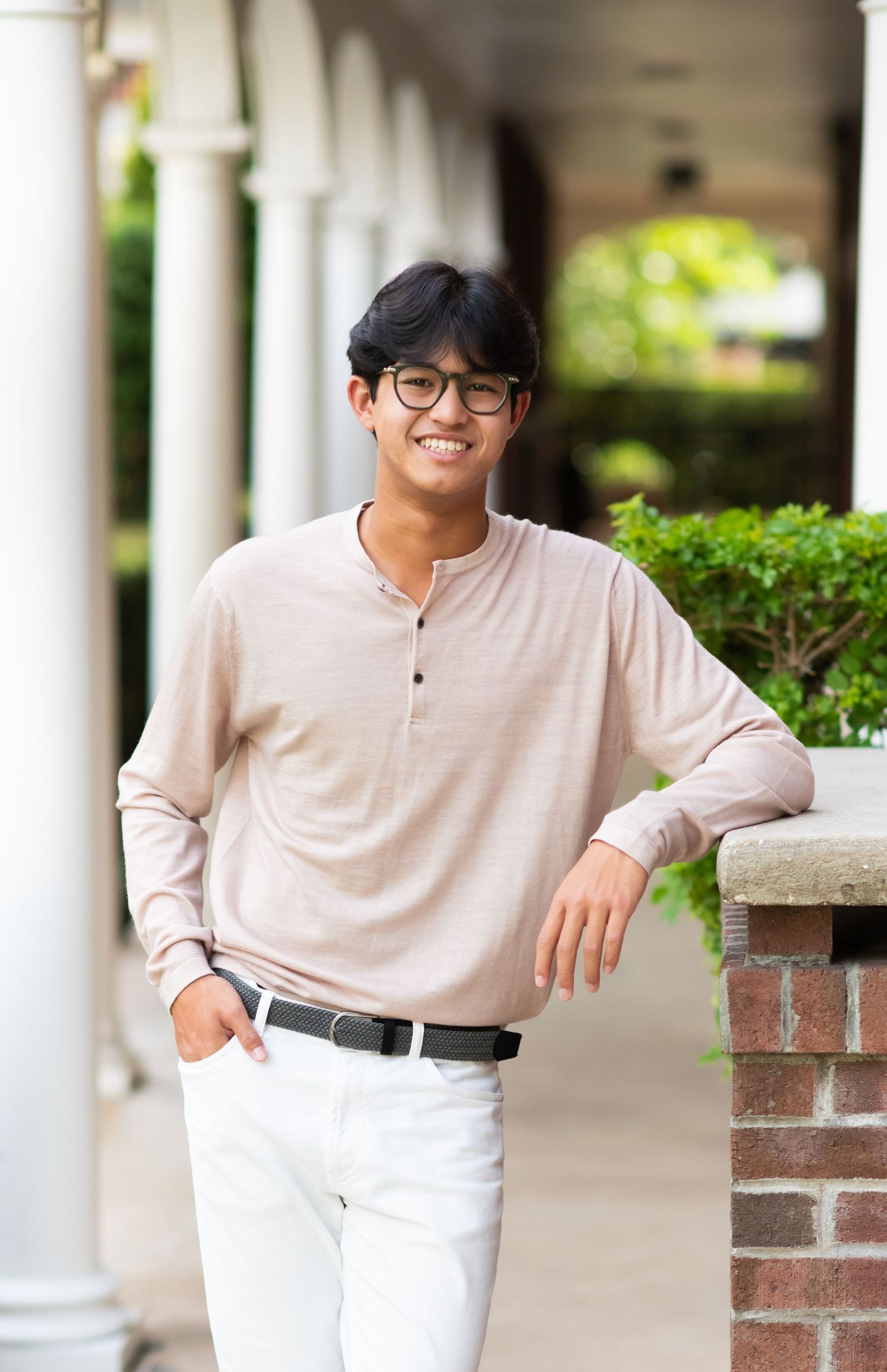 Smiling young man in beige henley leaning on brick pillar outdoors.