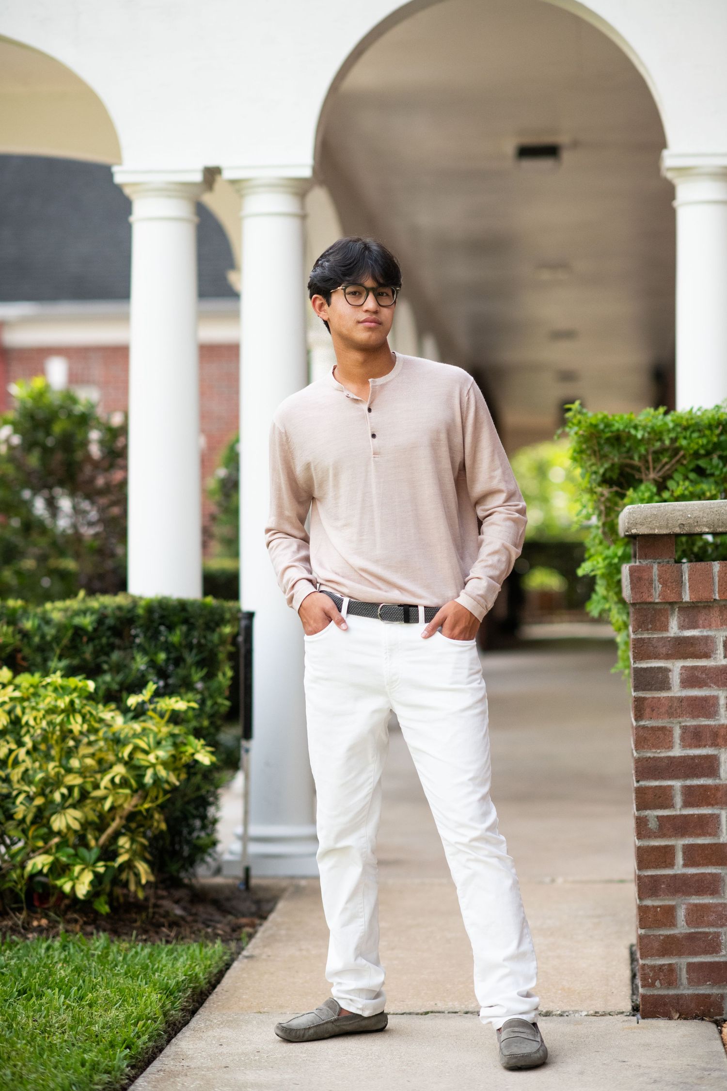 Young Asian man in beige henley and white pants standing by brick column.